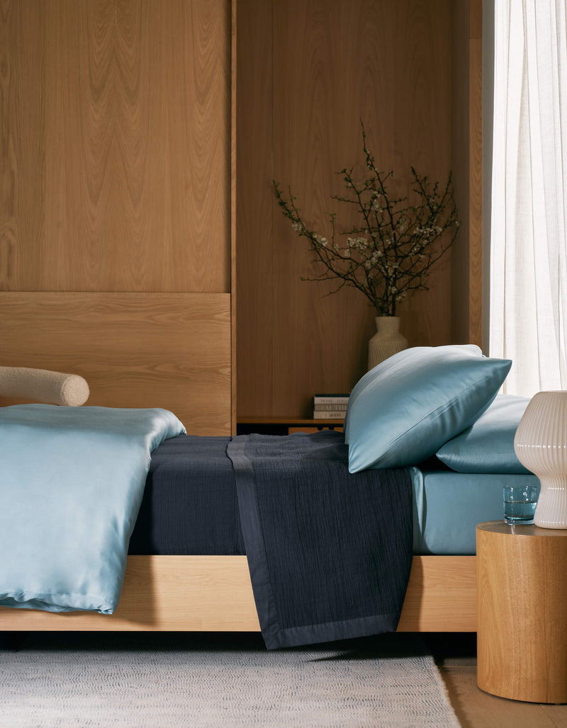 A modern bedroom features a light wood bed frame, blue satin bedding, and the Cozy Earth Waterfront Grid Matelasse Blanket in dark blue, with a wooden nightstand, ribbed white lamp, glass of water, and vase against wood-paneled walls and sheer curtains.