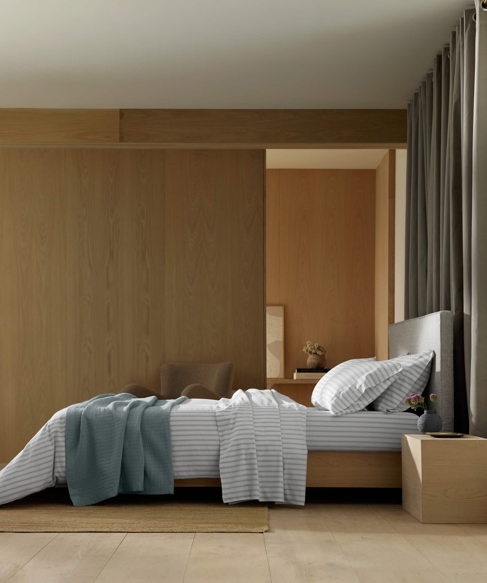 A modern bedroom with a neatly made bed featuring the Cozy Earth Waterfront Grid Matelasse Blanket, gray and white striped bedding, a blue throw, light wood headboard, bedside table with a plant, and neutral tones with wooden accents.