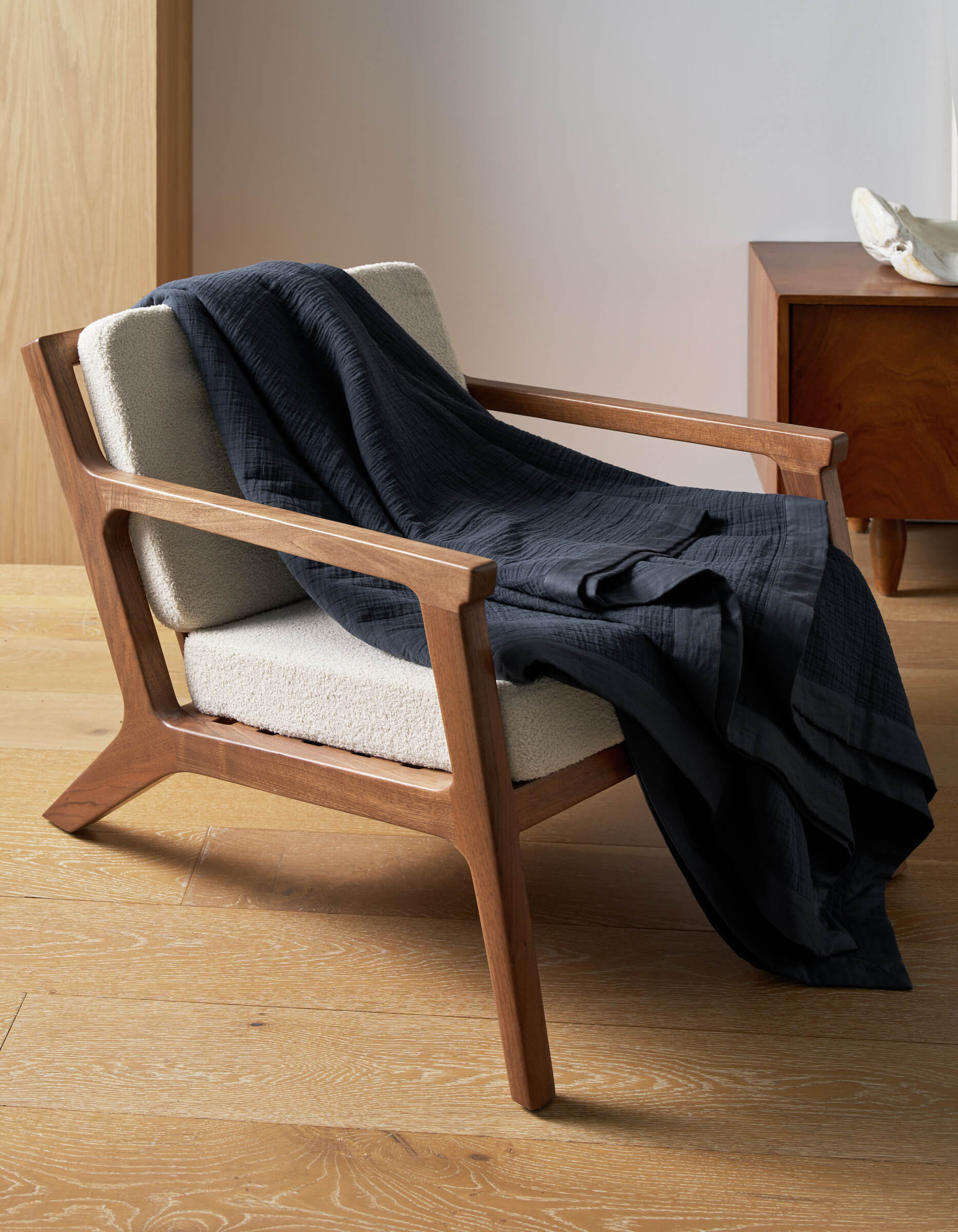 A wooden armchair with cream upholstery sits on a light wood floor, draped with a Cozy Earth Waterfront Grid Matelasse Blanket. A wooden side table and decorative object appear in the background.