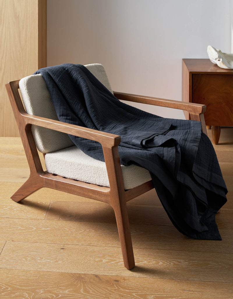 A wooden armchair with cream upholstery sits on a light wood floor, draped with a Cozy Earth Waterfront Grid Matelasse Blanket. A wooden side table and decorative object appear in the background.