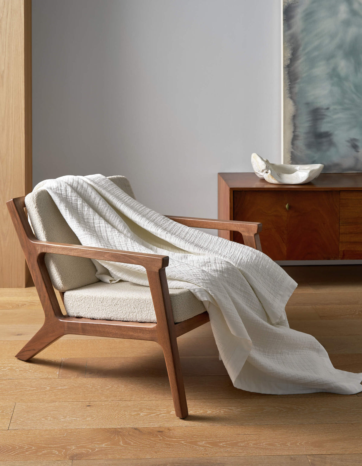 A wooden armchair with cream upholstery sits on a light wood floor, draped with the Cozy Earth Waterfront Grid Matelasse Blanket. In the background, there’s a wooden sideboard with a white bowl and large abstract artwork on the wall.|Color:Sea Salt