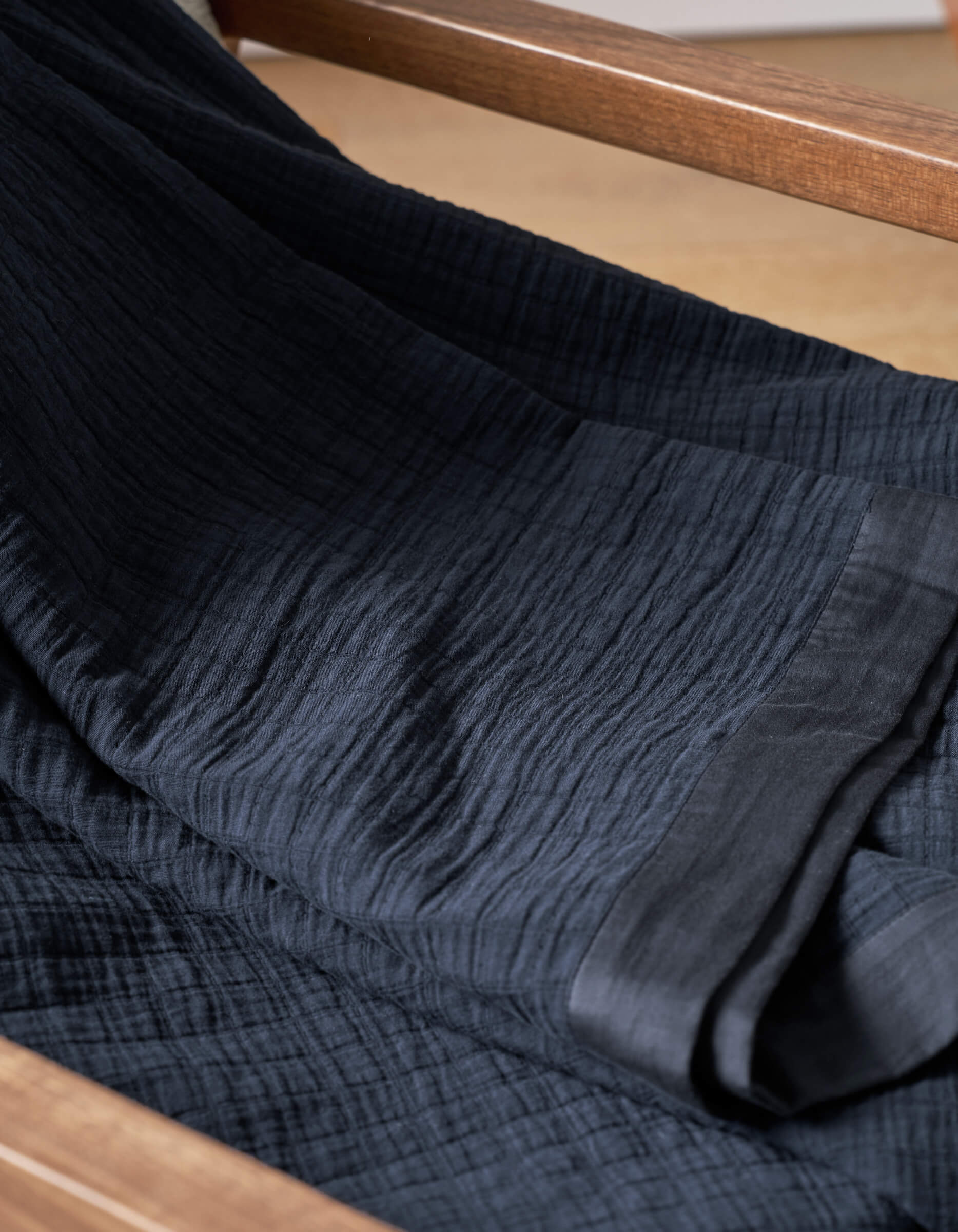 A close-up of Cozy Earth's Waterfront Grid Matelasse Blanket in dark navy, draped over wood, highlights its textured fabric with fine crinkles and a smooth edge.