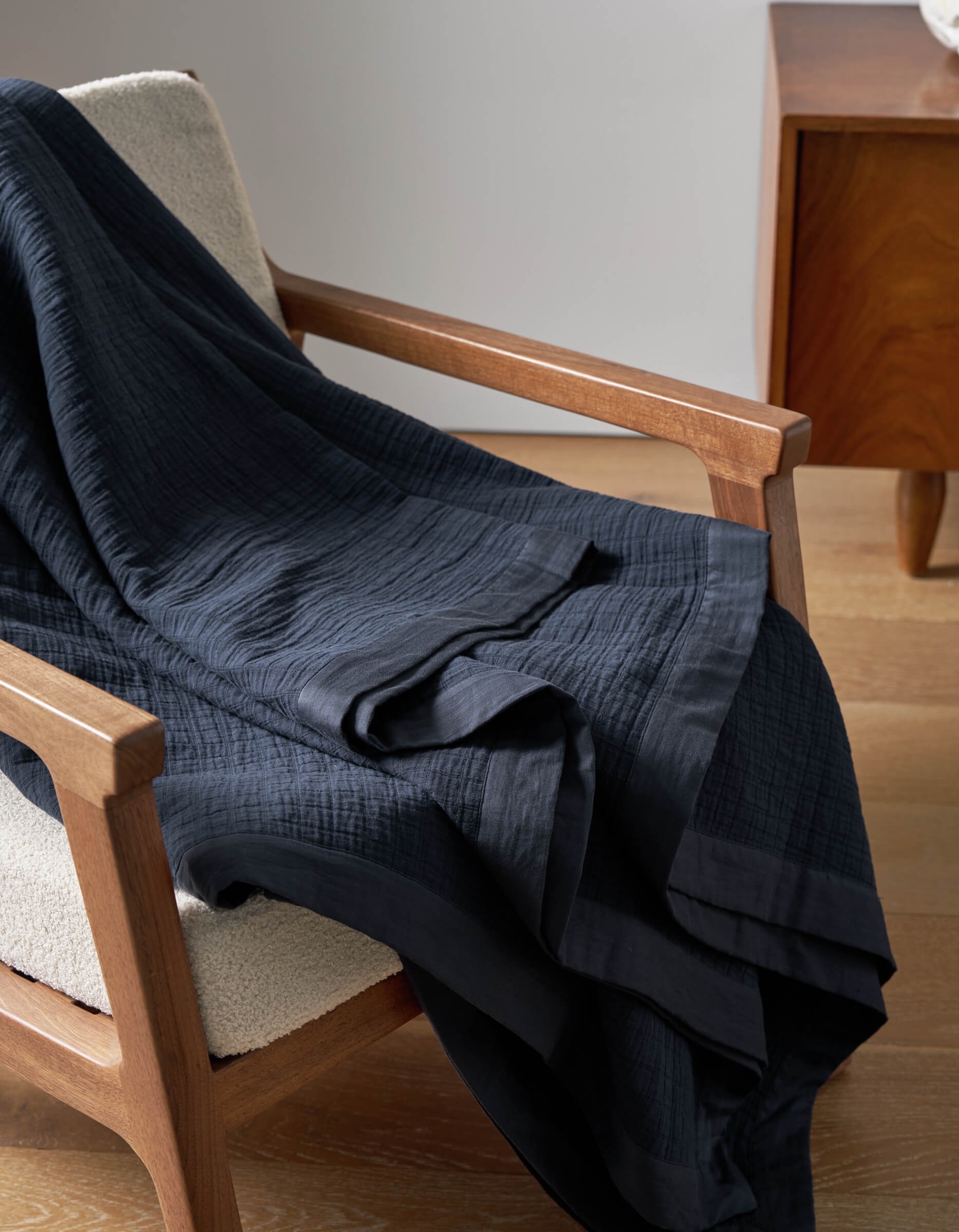 The Cozy Earth Waterfront Grid Matelasse Blanket in dark navy blue is draped over a light upholstered wooden armchair beside a wooden side table on a light wood floor.