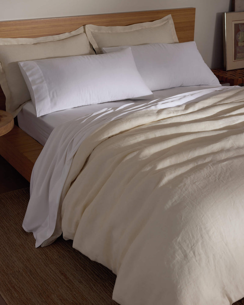 A bed features the Cozy Earth Linen Duvet Cover, white pillows, and a wooden headboard. A bedside table adds warmth to the softly lit room, creating a cozy and inviting atmosphere.