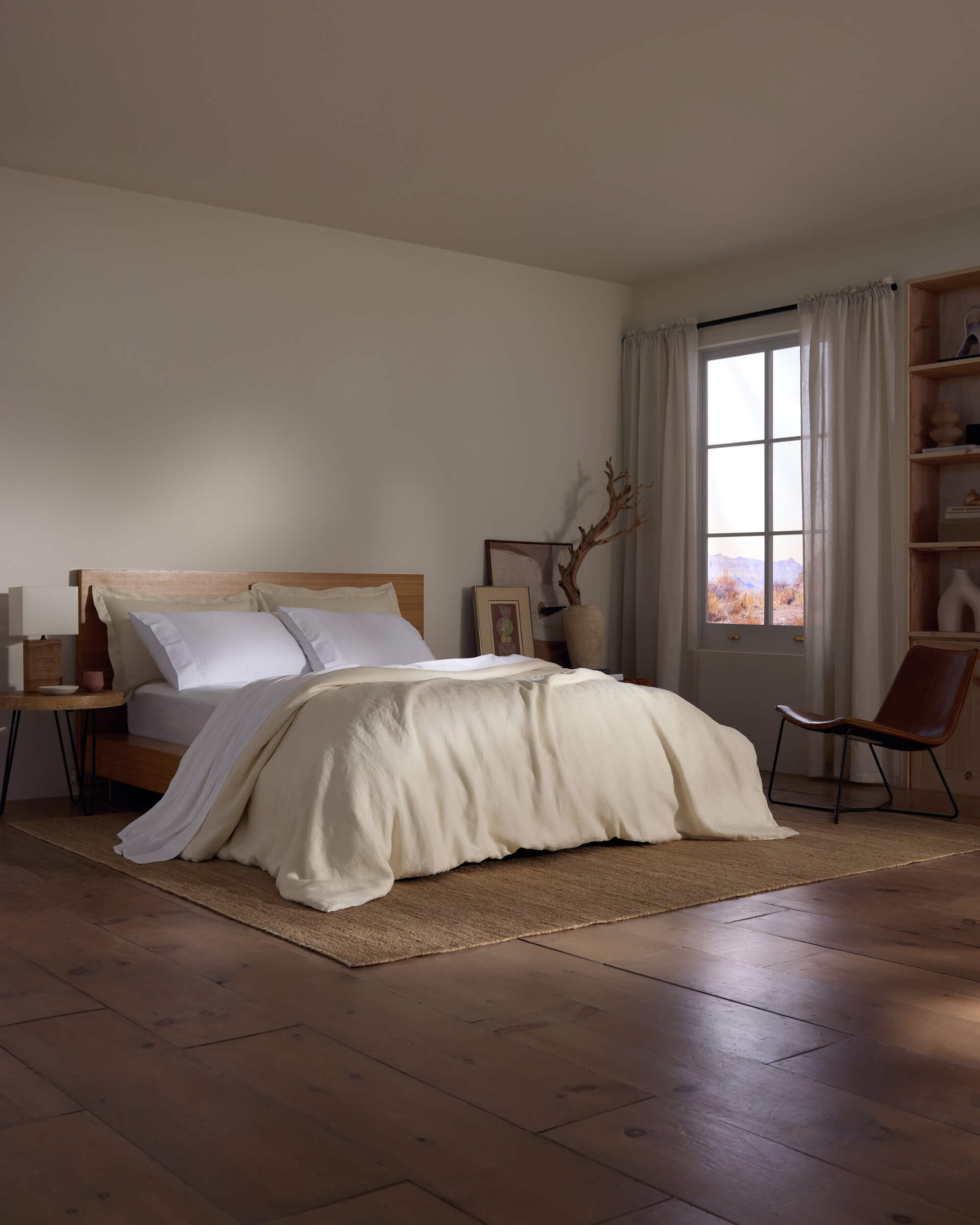 A minimal bedroom with wooden floors, a Cozy Earth Linen Duvet Cover on white bedding, a wooden bed frame, bedside table, window with sheer curtains, and a chair by shelves and decor on the right wall.