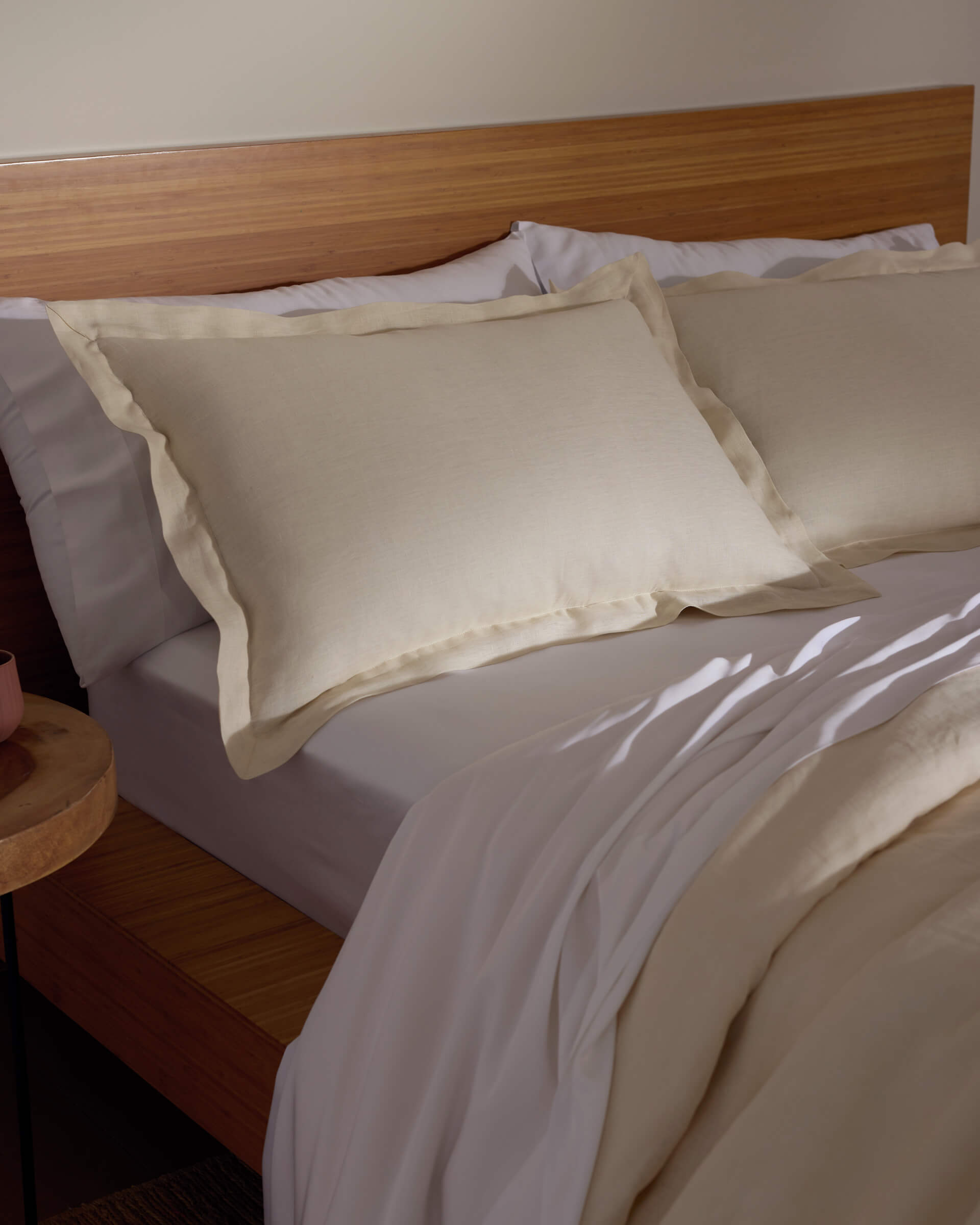 A bed featuring Cozy Earth's Linen Shams in cream, paired with a matching comforter, sits against a wooden headboard and nightstand, all bathed in soft natural light.