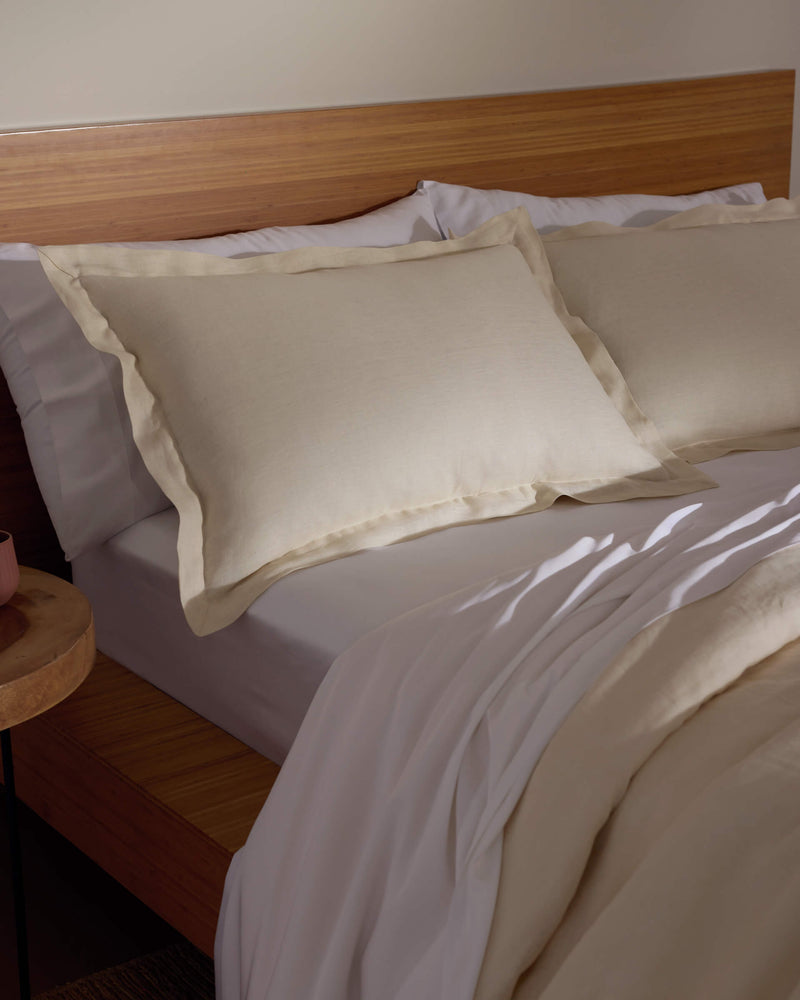A bed featuring Cozy Earth's Linen Shams in cream, paired with a matching comforter, sits against a wooden headboard and nightstand, all bathed in soft natural light.