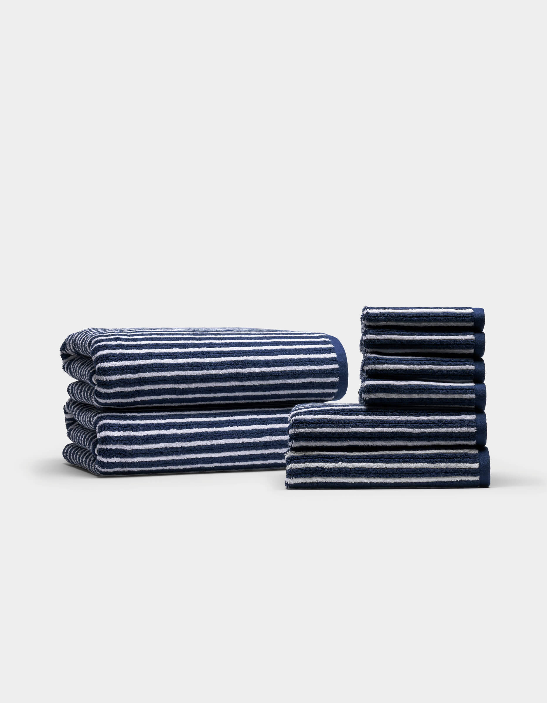Cozy Earth's Ribbed Terry Bath Towel Set, featuring neatly folded navy blue and white striped towels, is stacked in two organized piles against a plain light background. The towels are arranged by size, creating even and orderly stacks.|Color:Obsidian Stripe