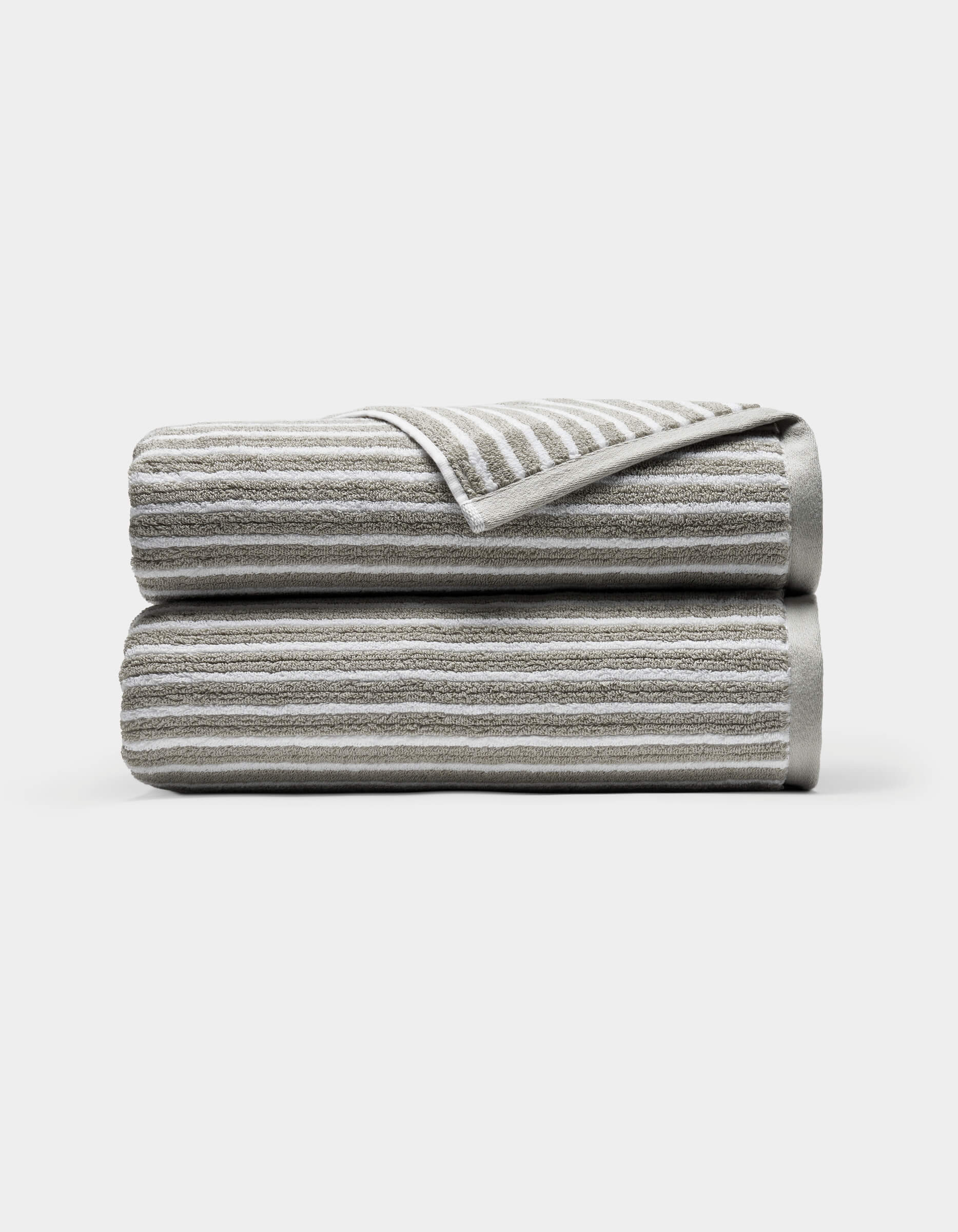 Two neatly folded Ribbed Terry Bath Towels by Cozy Earth, light gray with horizontal white stripes, stacked atop each other against a plain white background. A corner of the top towel is turned over, revealing the underside.|Color:Dew Stripe