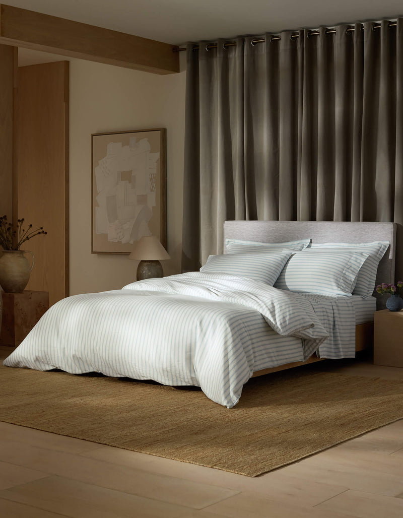 A neatly made bed features the Cozy Earth Soft-Wash Cotton Bedding Core Bundle in light gray and white stripes, set atop a woven rug. Gray curtains, abstract wall art, and minimalist decor create a cozy, modern bedroom ambiance.