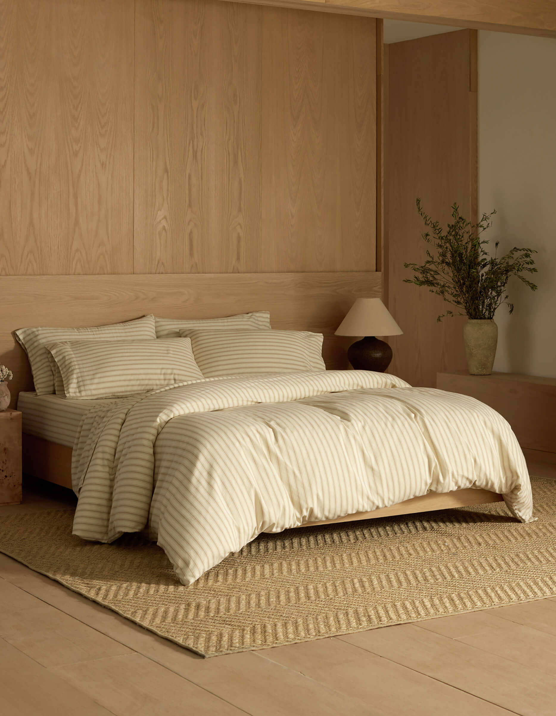 A minimalist bedroom features Cozy Earth's Soft-Wash Cotton Bedding Core Bundle in cream and beige stripes on the bed, paired with light wood walls and floor, a textured rug, bedside lamp, and a vase with greenery on a low cabinet.