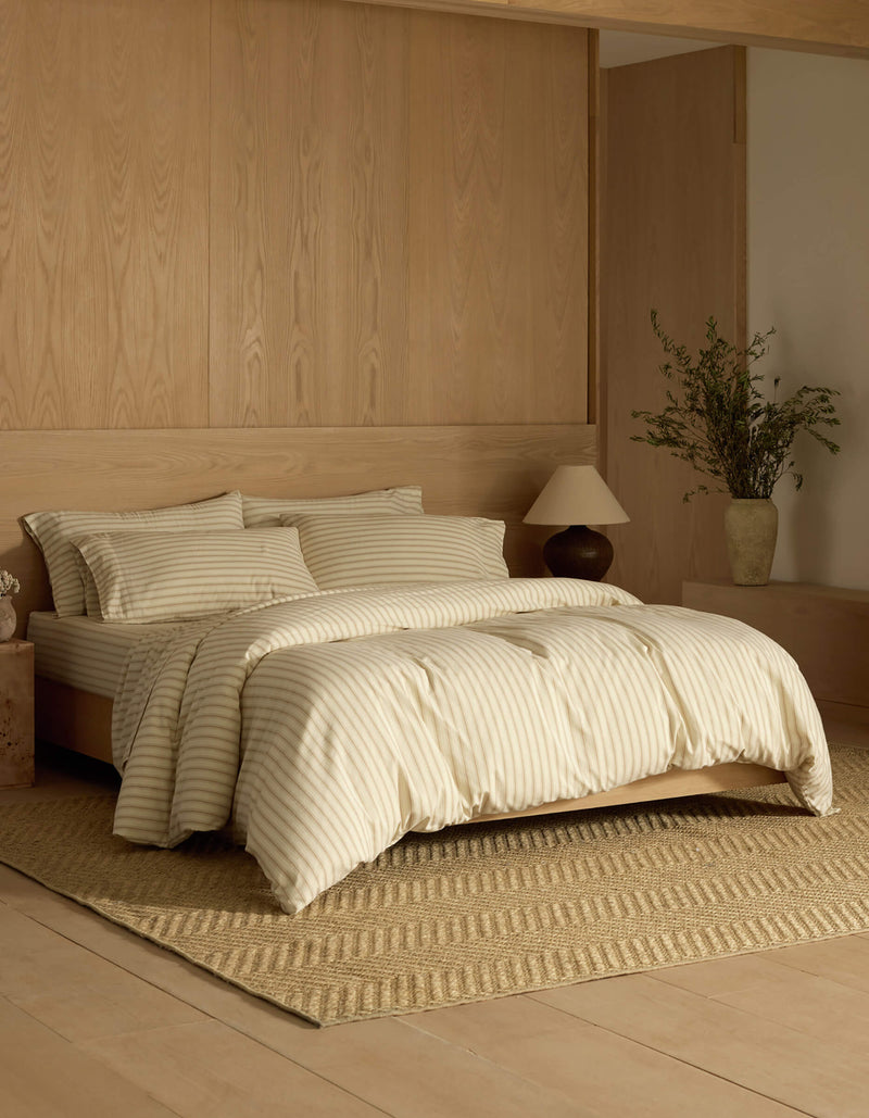 A minimalist bedroom features Cozy Earth's Soft-Wash Cotton Bedding Core Bundle in cream and beige stripes on the bed, paired with light wood walls and floor, a textured rug, bedside lamp, and a vase with greenery on a low cabinet.