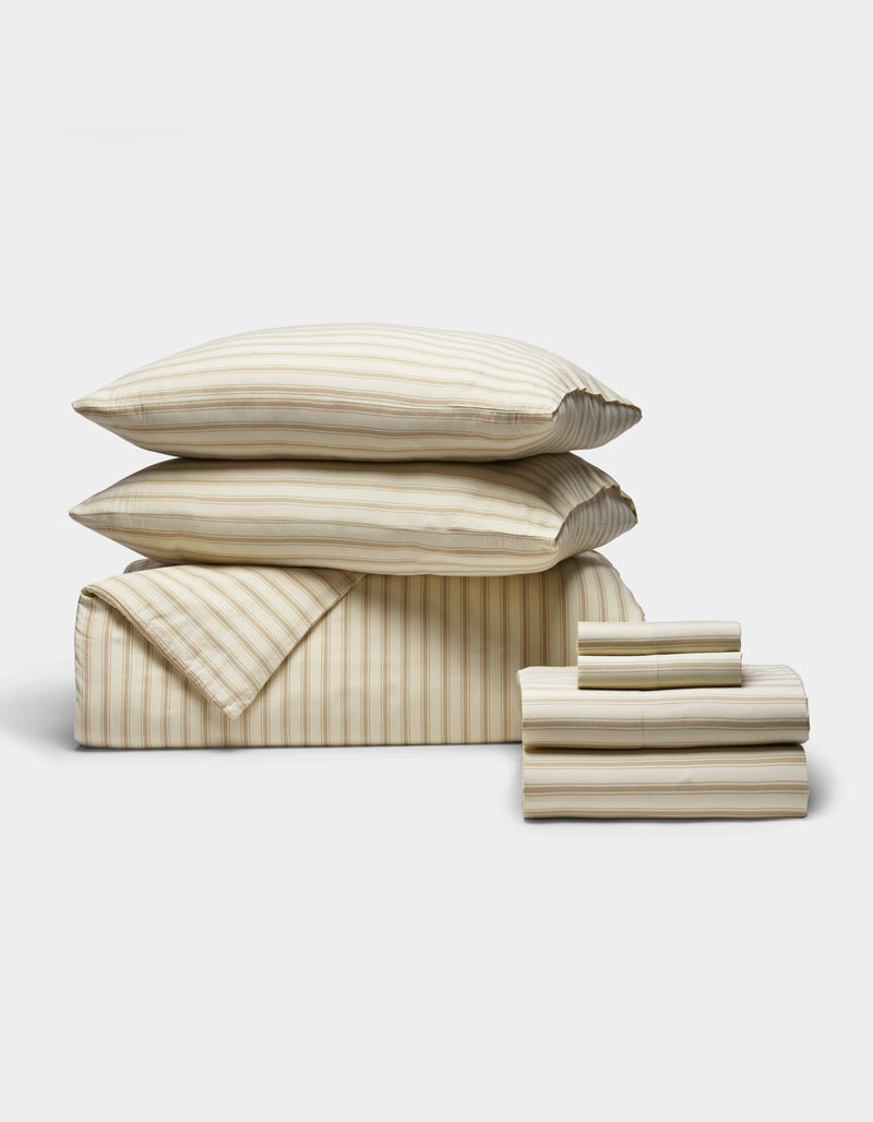 The Soft-Wash Cotton Bedding Core Bundle by Cozy Earth features cream and beige striped bedding with two stacked pillows, a folded comforter, and a neatly folded sheet stack, all displayed against a plain white background.