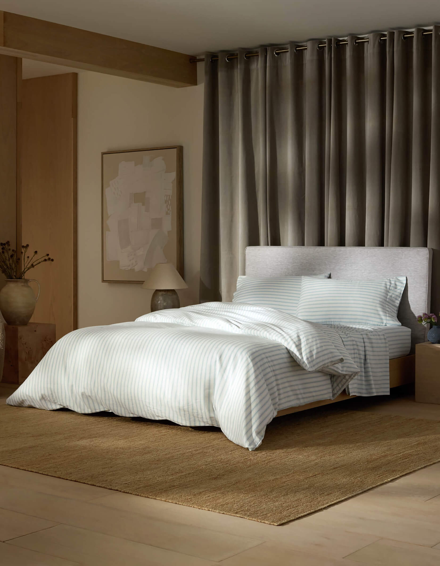 A modern bedroom with a neatly made bed featuring Cozy Earth's Soft-Wash Cotton Duvet Cover, a light headboard, brown textured rug, gray floor-length curtains, bedside table with a lamp and vase, and abstract wall art.