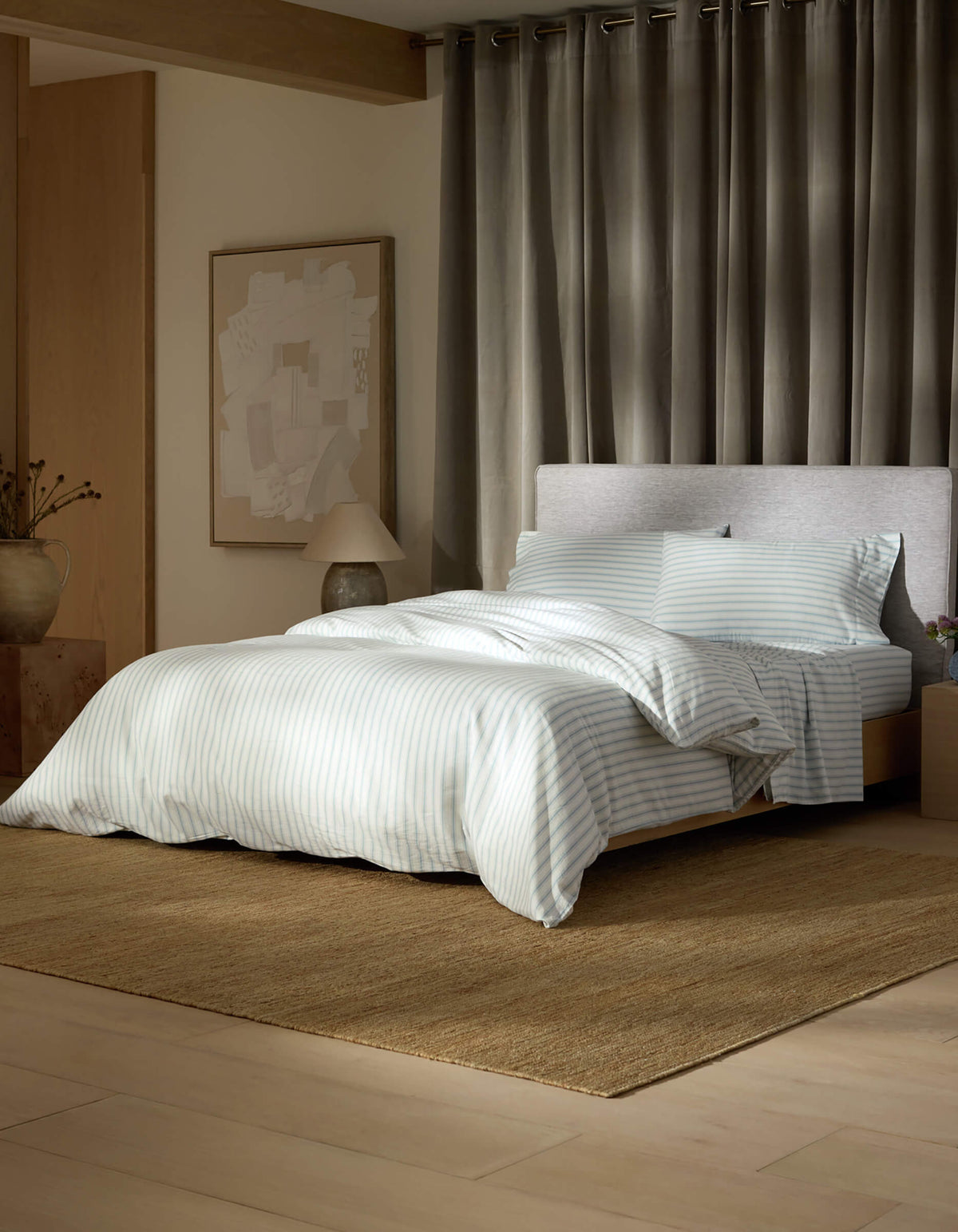 A modern bedroom features the Soft-Wash Cotton Bedding Deluxe Bundle by Cozy Earth in light blue and white stripes, on a brown rug with grey curtains, an abstract art piece, a lamp, and a decorative vase on the side table.|Color:River and Sea Salt Stripe