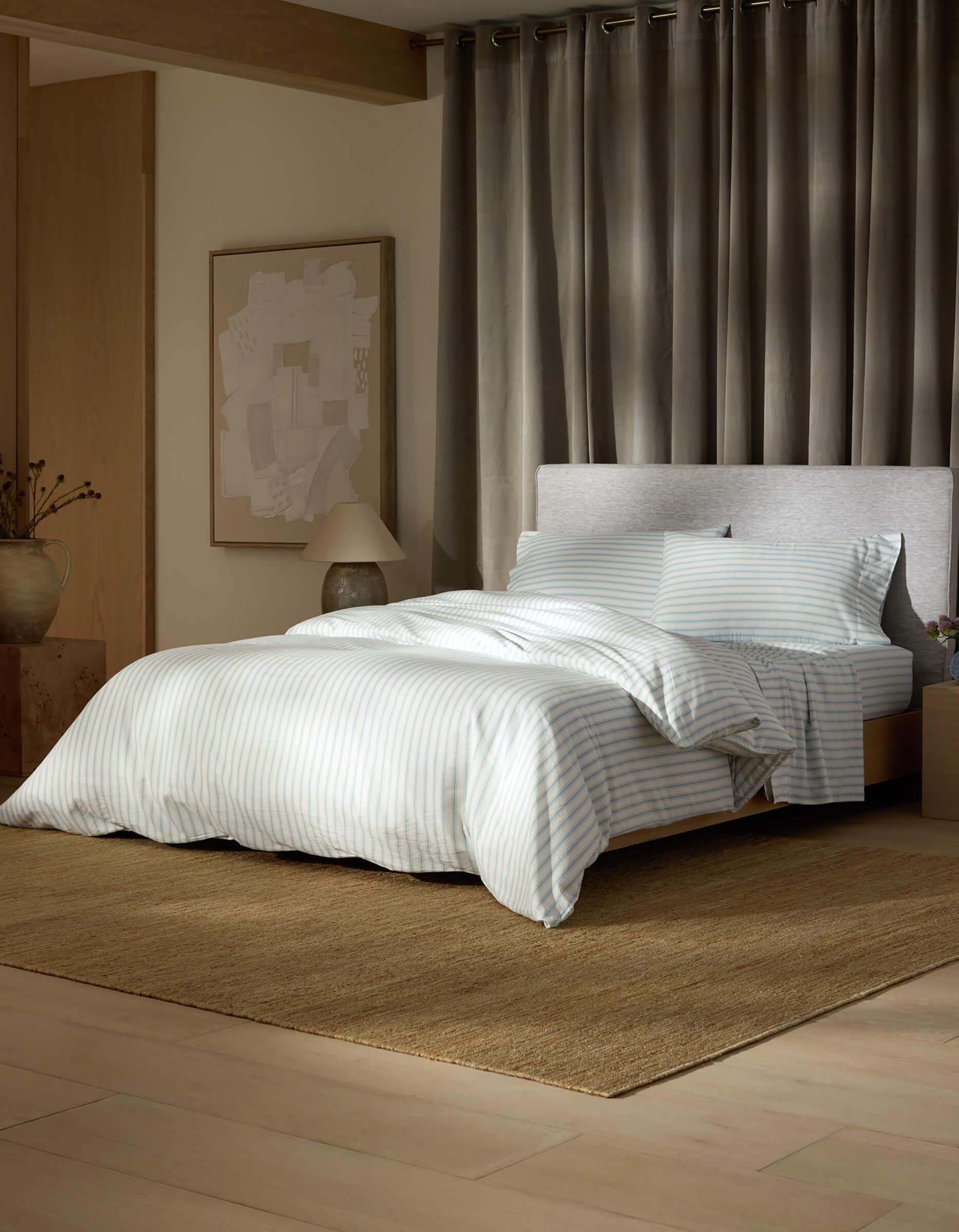 A modern bedroom features the Soft-Wash Cotton Bedding Deluxe Bundle by Cozy Earth in light blue and white stripes, on a brown rug with grey curtains, an abstract art piece, a lamp, and a decorative vase on the side table.