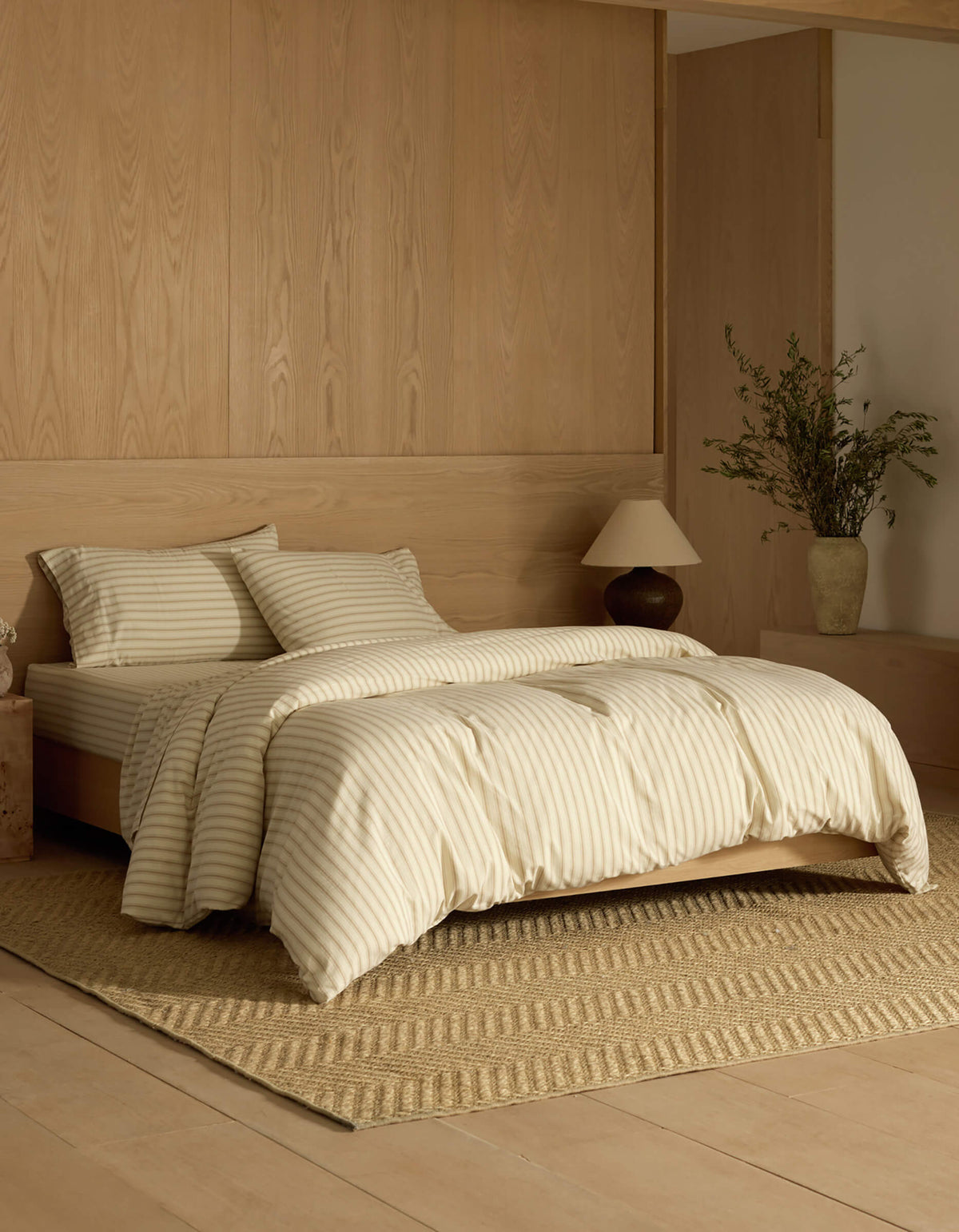 A neatly made bed with Cozy Earth's Soft-Wash Cotton Bedding Deluxe Bundle sits on a woven rug in a room with light wood walls and flooring. A nightstand with a lamp and vase of greenery stands beside the bed.|Color:Coconut and Sandalwood Stripe