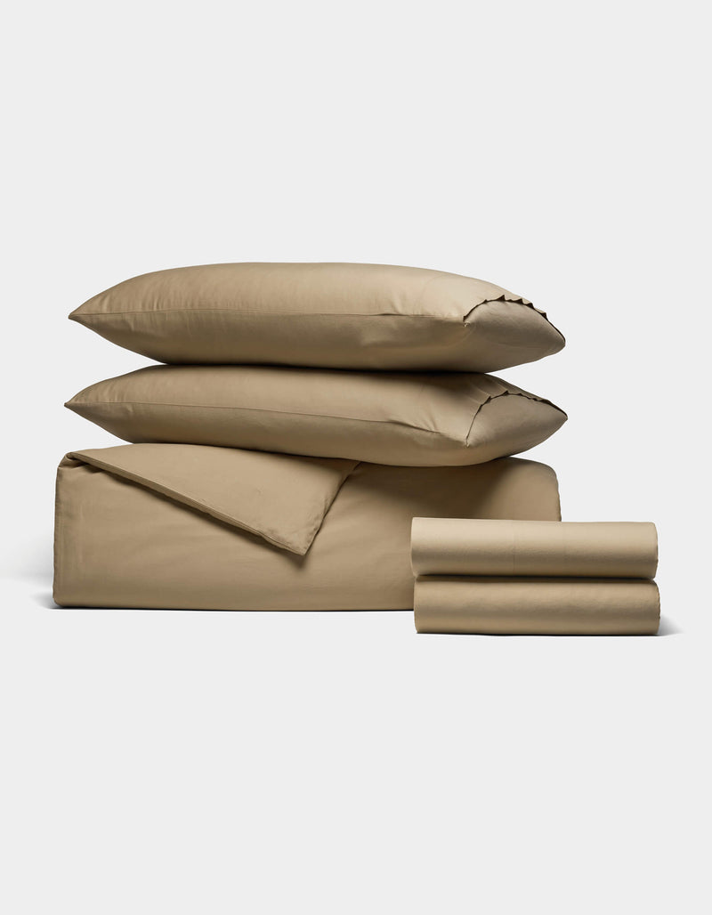 The Cozy Earth Soft-Wash Cotton Bedding Deluxe Bundle features two pillows, a folded duvet, and two stacks of tan sheets, all neatly arranged on a white background.