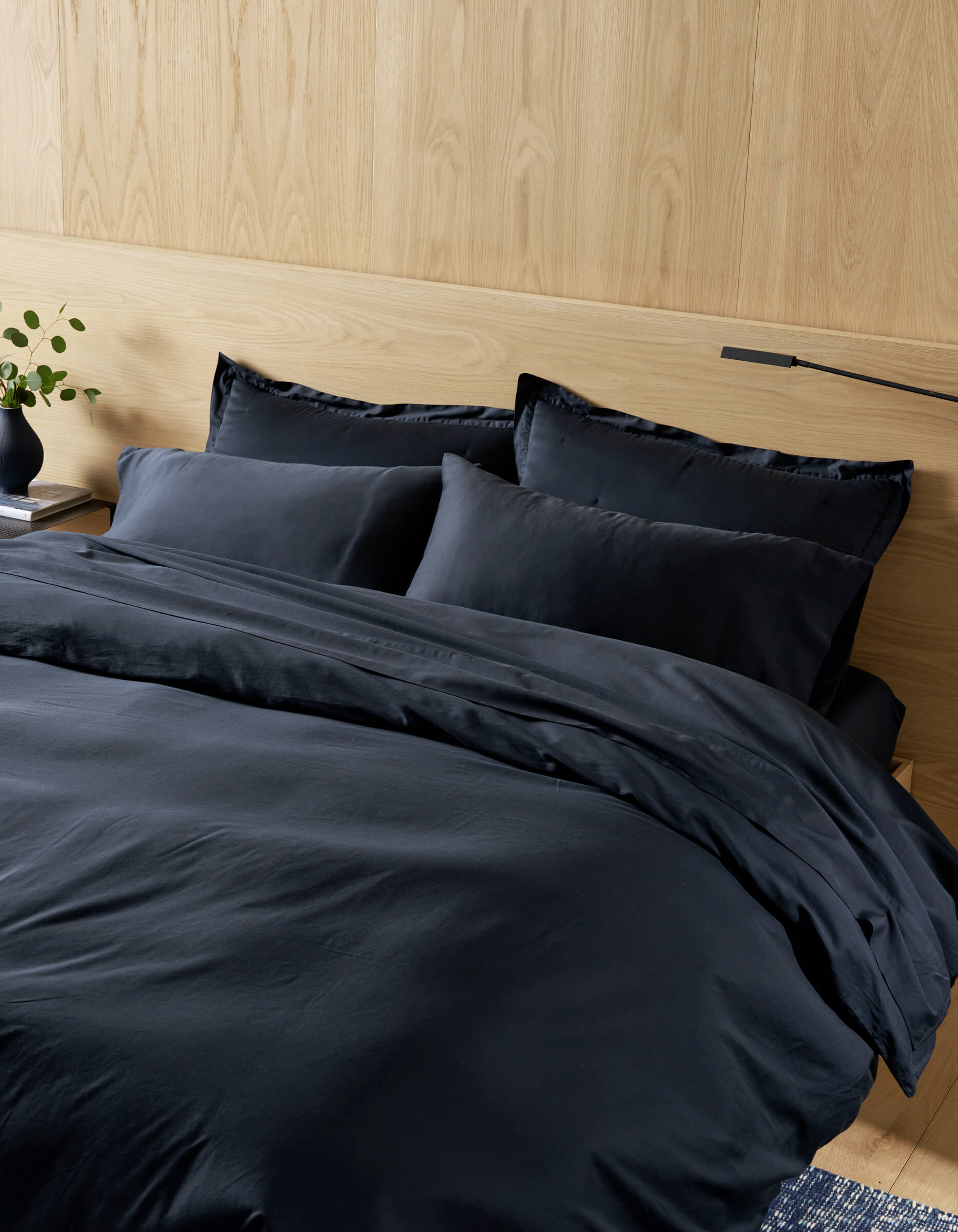 A neatly made bed features the Cozy Earth Soft-Wash Cotton Duvet Cover in dark navy with matching pillows, set against a light wooden headboard. A nightstand on the left holds a black vase with green leaves.