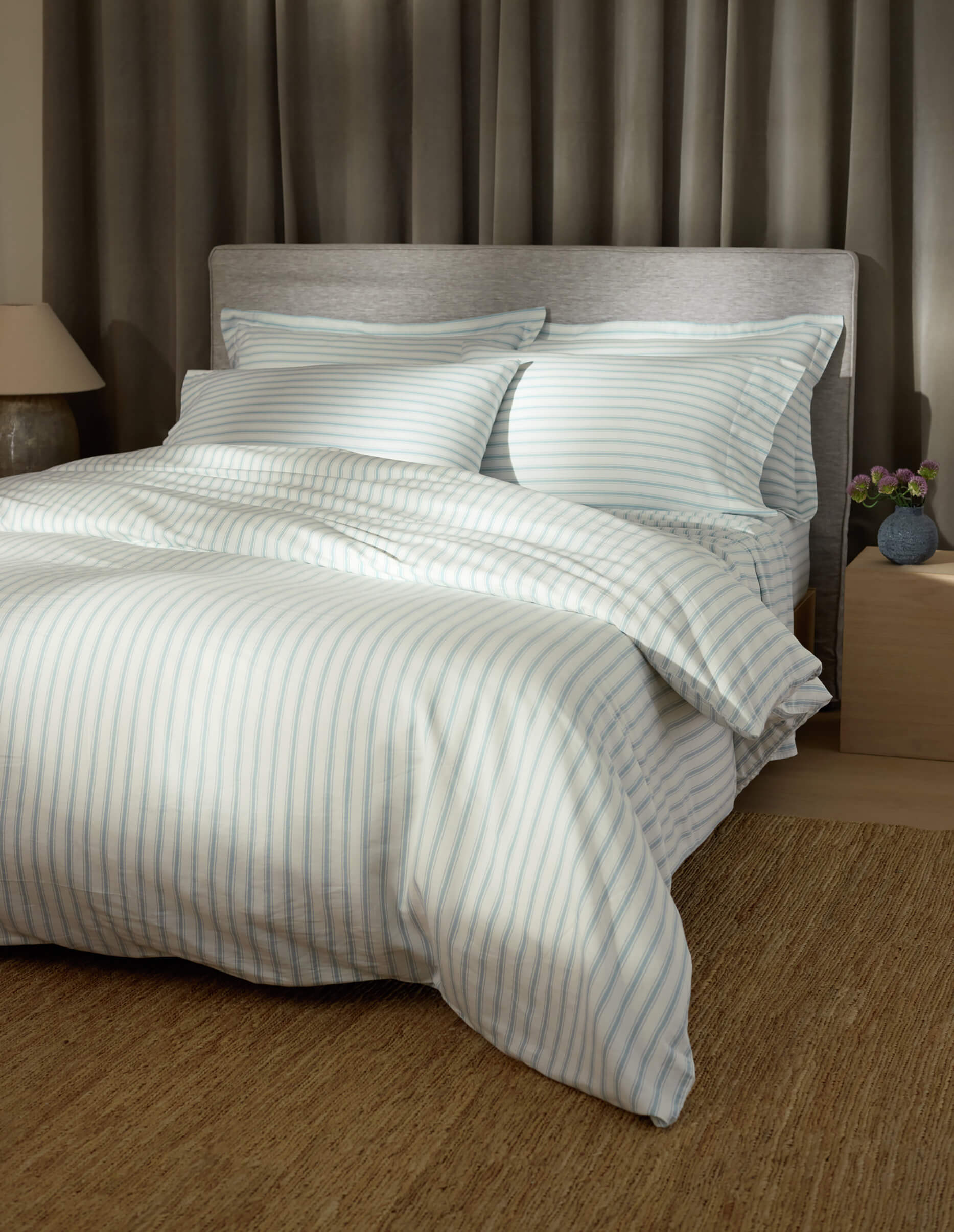 A cozy room featuring Cozy Earth's Soft-Wash Cotton Duvet Cover in light blue and white stripes, two pillows, a gray upholstered headboard, beige curtains, a bedside table with a lamp, and a small vase of flowers.