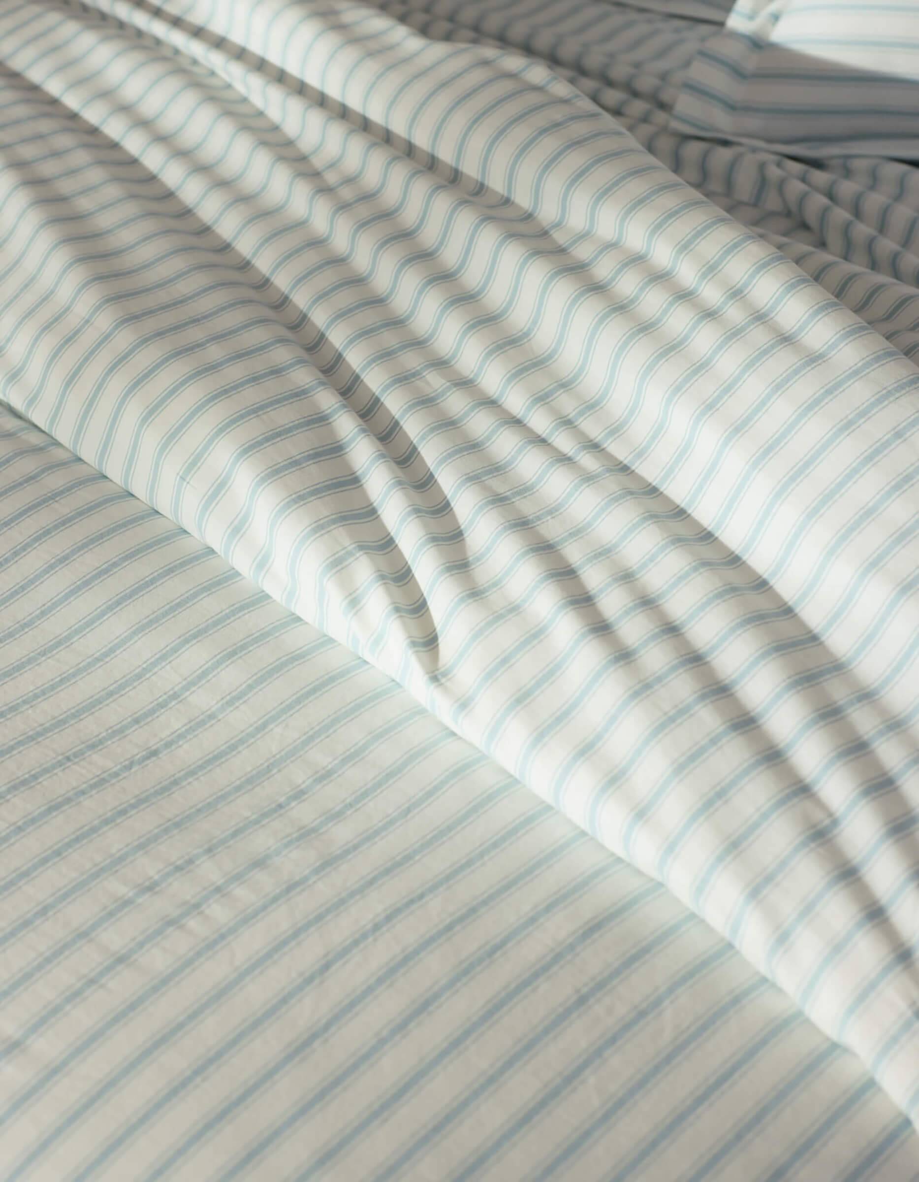 Close-up of a bed featuring the Cozy Earth Soft-Wash Cotton Duvet Cover in white and light blue stripes, gently wrinkled and illuminated by natural sunlight.