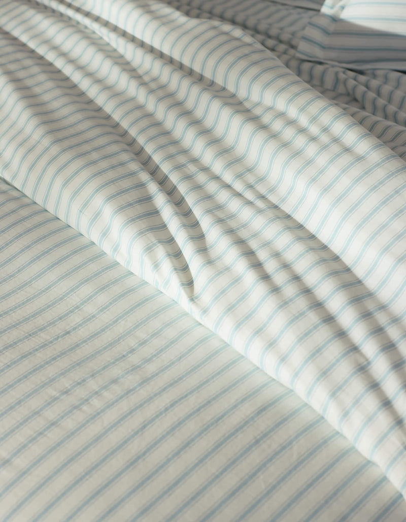 Close-up of a bed featuring the Cozy Earth Soft-Wash Cotton Duvet Cover in white and light blue stripes, gently wrinkled and illuminated by natural sunlight.