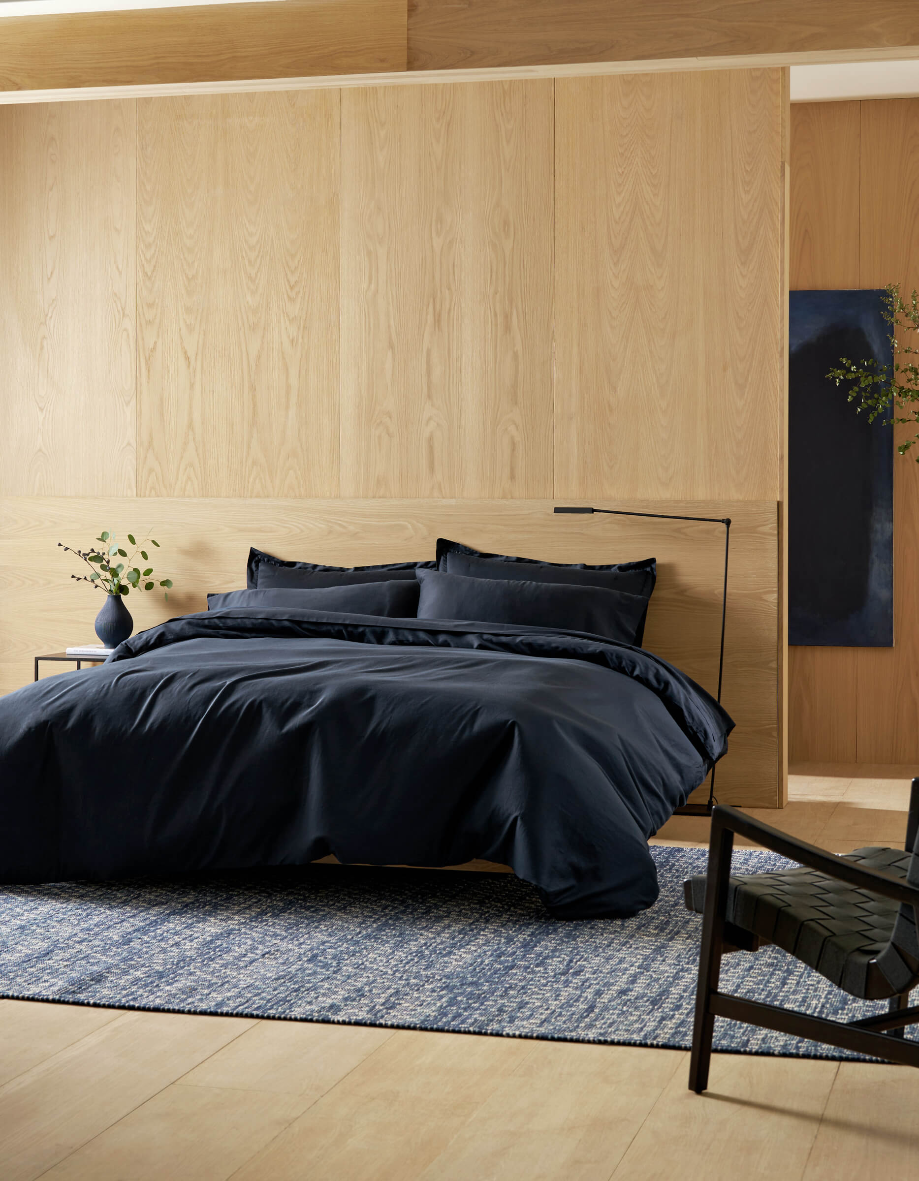 A modern bedroom features the Cozy Earth Soft-Wash Cotton Duvet Cover in black, with pillows, light wood walls and floors, a blue patterned rug, a black chair, and a small bedside table displaying a plant in a vase.