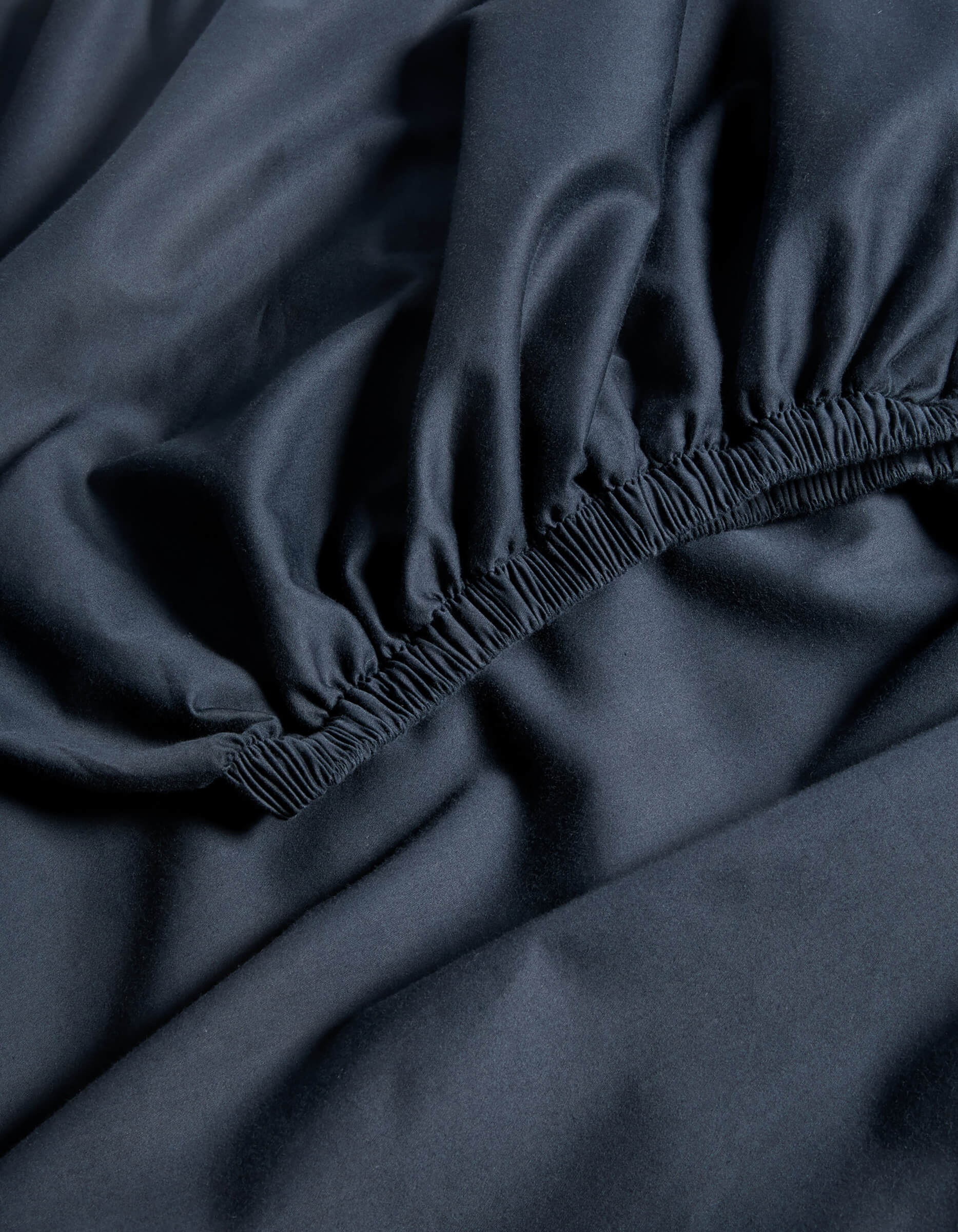 Close-up of Cozy Earth's Soft-Wash Cotton Fitted Sheet in a dark shade, showing the elasticized edges as it rests on a flat surface.