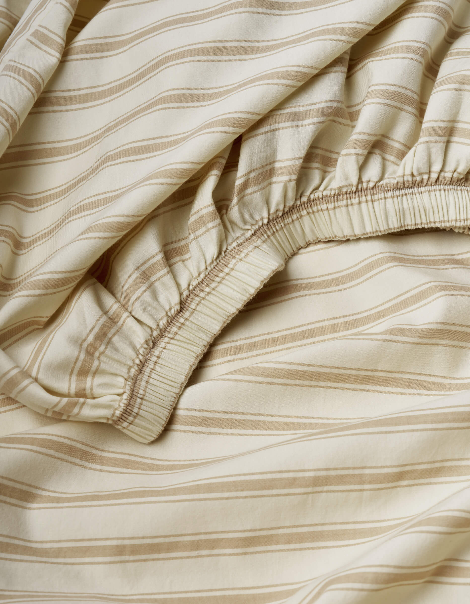 The Soft-Wash Cotton Fitted Sheet by Cozy Earth features a beige color with light brown horizontal stripes and an elastic edge, showcasing its gathered fabric detail, texture, and pattern.