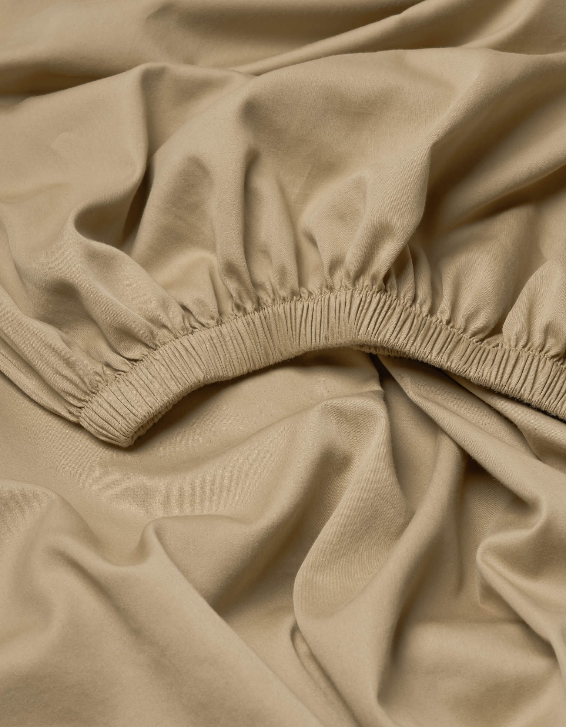 The Cozy Earth Soft-Wash Cotton Fitted Sheet features beige fabric with elasticated ruching, forming gathered folds for a soft, textured look. The smooth, slightly shiny material is arranged in loose, overlapping layers.