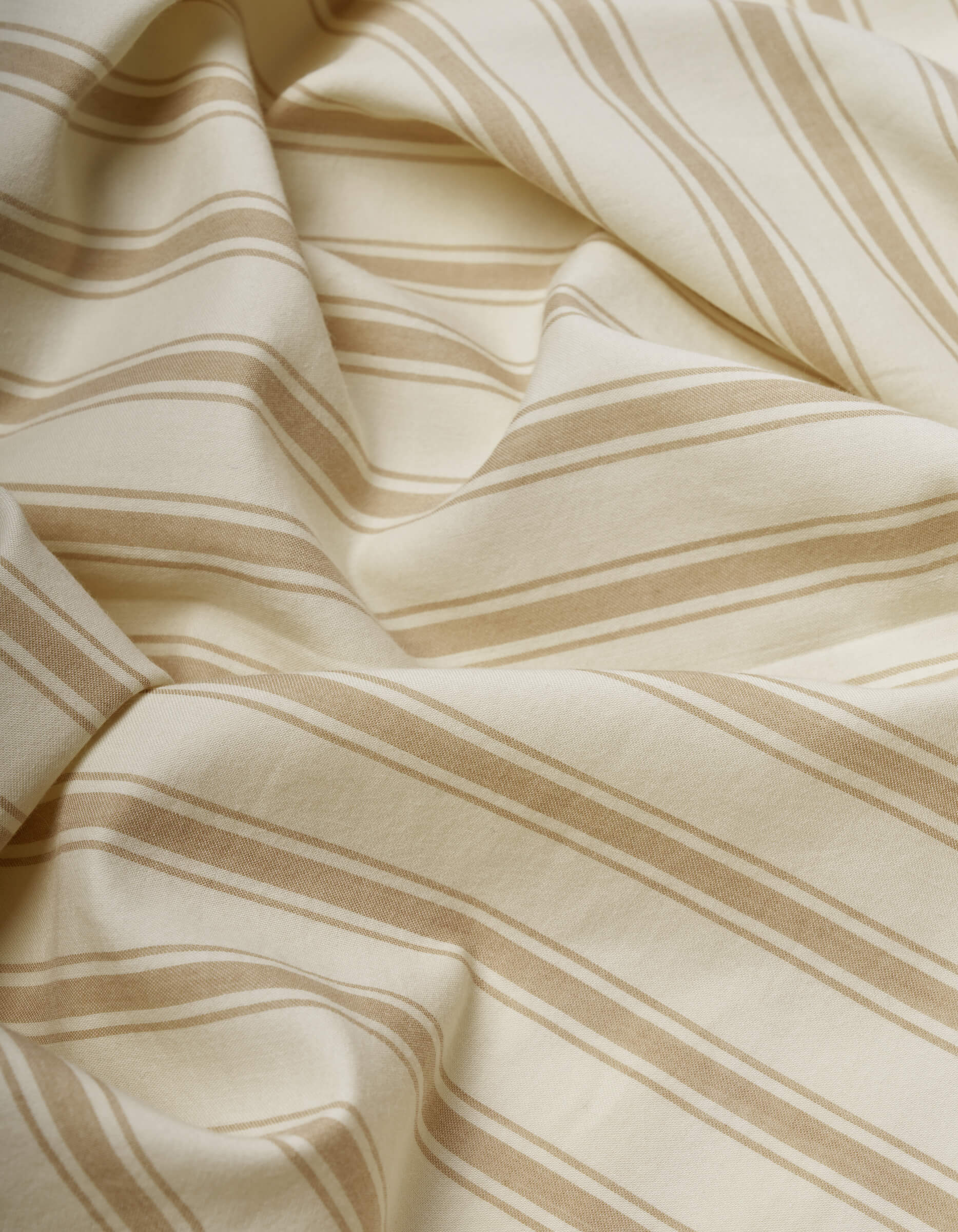 The Cozy Earth Soft-Wash Cotton Flat Sheet features cream-colored fabric with light brown diagonal stripes, softly draped with gentle folds and subtle shadows across its slightly wrinkled surface.