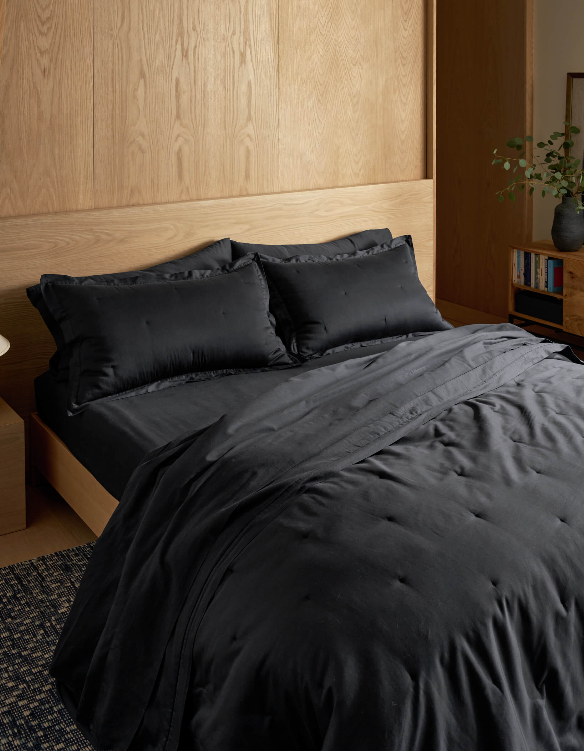 A modern, minimalist room features a neatly made bed with Cozy Earth’s Soft-Wash Cotton Quilt in black, set against a light wood headboard and wall. In the corner, a bookshelf holds books and a vase with greenery.