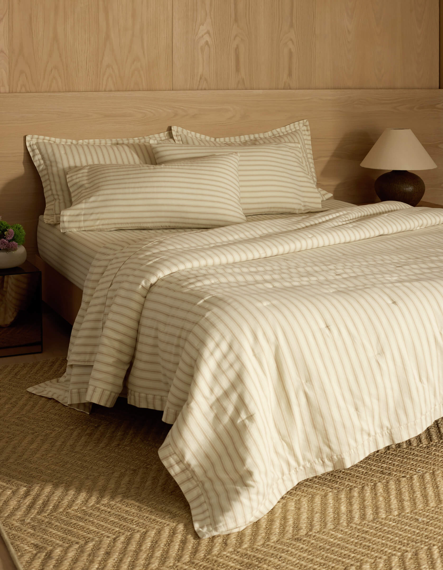 A neatly made bed features the Cozy Earth Soft-Wash Cotton Quilt in cream and beige stripes, with matching pillows and comforter. Set against a light wood-paneled wall, a bedside table holds a lamp and vase; a woven rug lies on the floor.