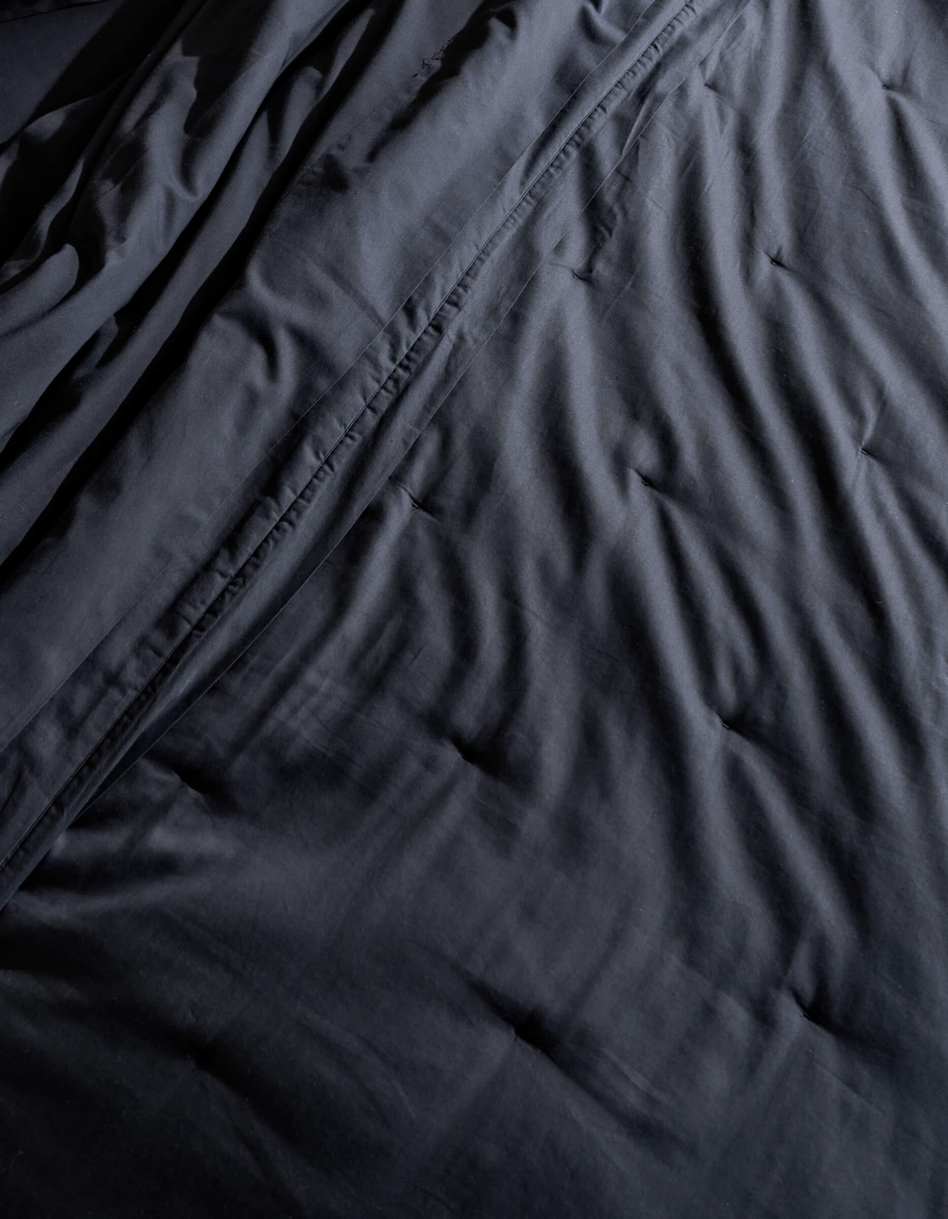 A close-up of Cozy Earth's Soft-Wash Cotton Quilt shows its dark, crumpled fabric with visible wrinkles and folds. The soft lighting highlights the quilt's subtle sheen and textured appearance.