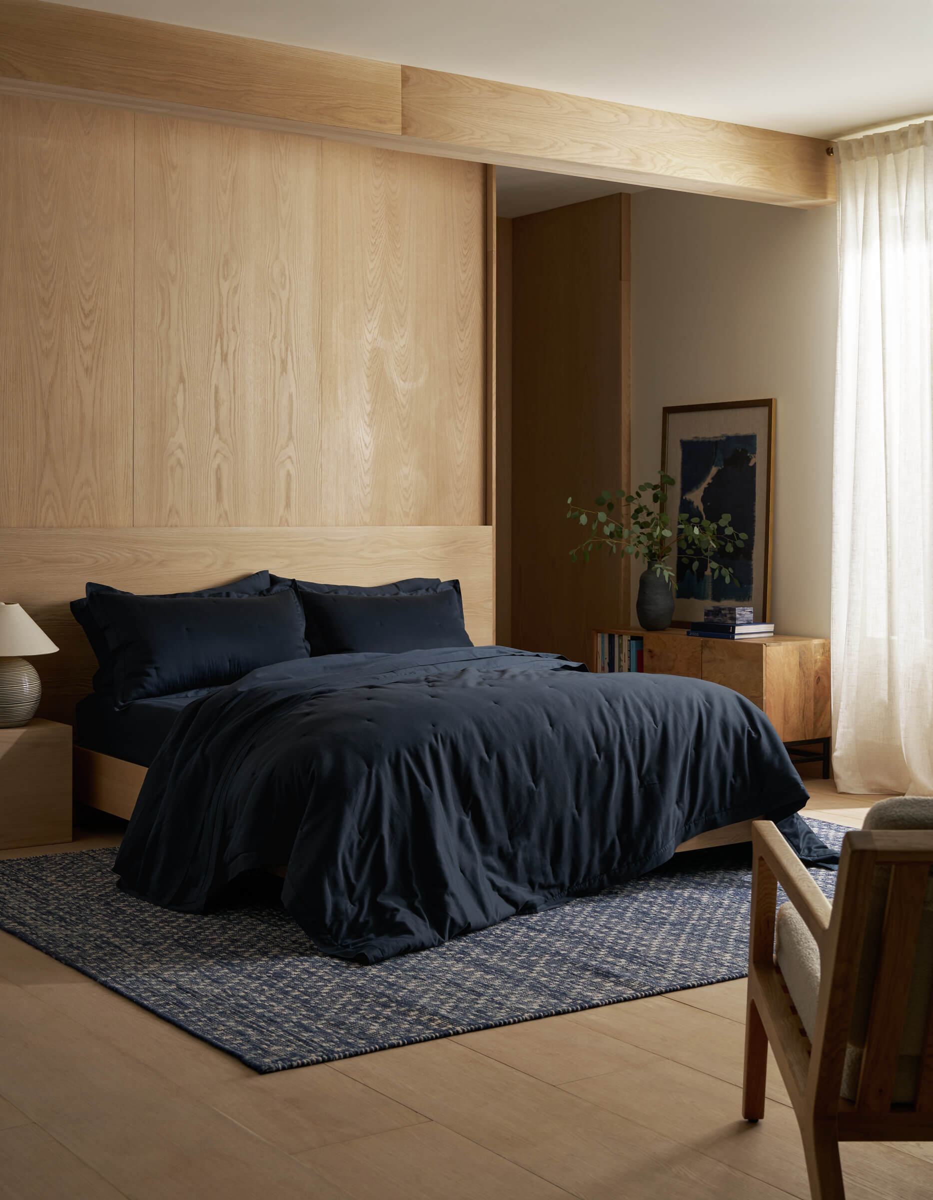A modern bedroom features light wood walls and floors, a bed dressed in Cozy Earth’s Soft-Wash Cotton Quilt Set, a blue patterned rug, wooden nightstand with lamp, sunlight through sheer curtains, artwork, and a plant.