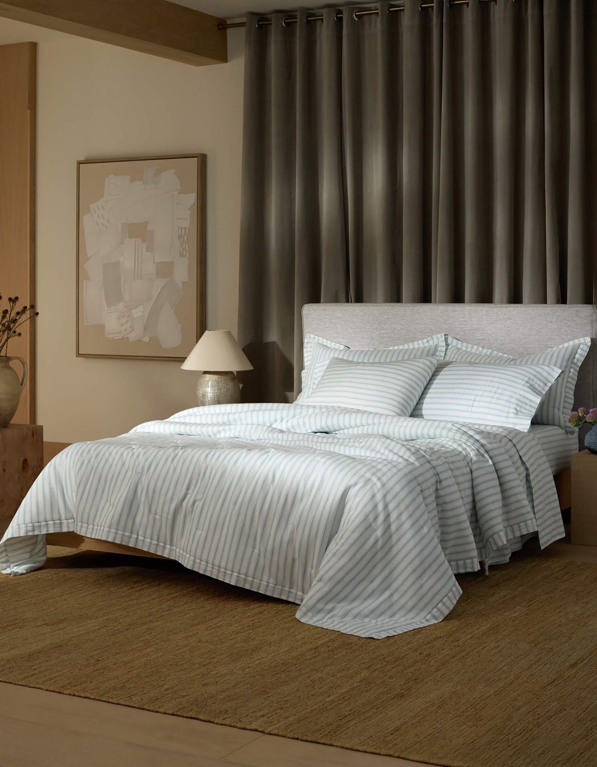 A modern bedroom features the Cozy Earth Soft-Wash Cotton Quilt in light blue and white stripes, set on a neatly made bed with a gray upholstered headboard, abstract wall art, bedside table with lamp and vase, gray curtains, and a woven rug.|Color:River and Sea Salt Stripe