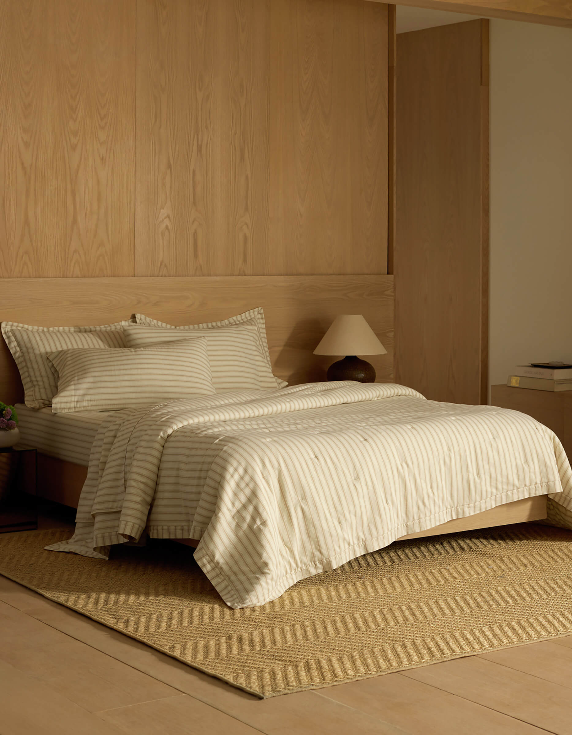 A cozy bedroom featuring Cozy Earth's Soft-Wash Cotton Quilt, cream and beige striped bedding, two pillows, a wooden headboard, a small bedside lamp, a woven rug, and minimalistic wooden furniture in neutral tones.