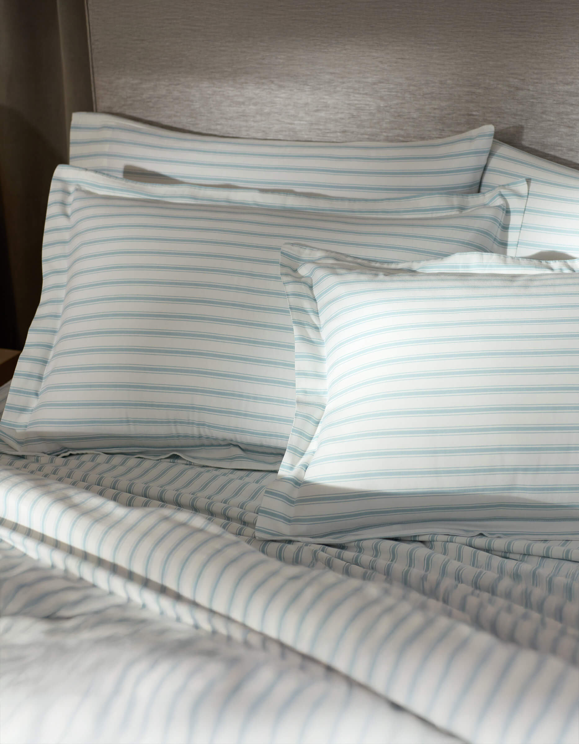 A bed featuring Cozy Earth Soft-Wash Cotton Shams in white and light blue stripes is set against a gray upholstered headboard, softly illuminated by sunlight.