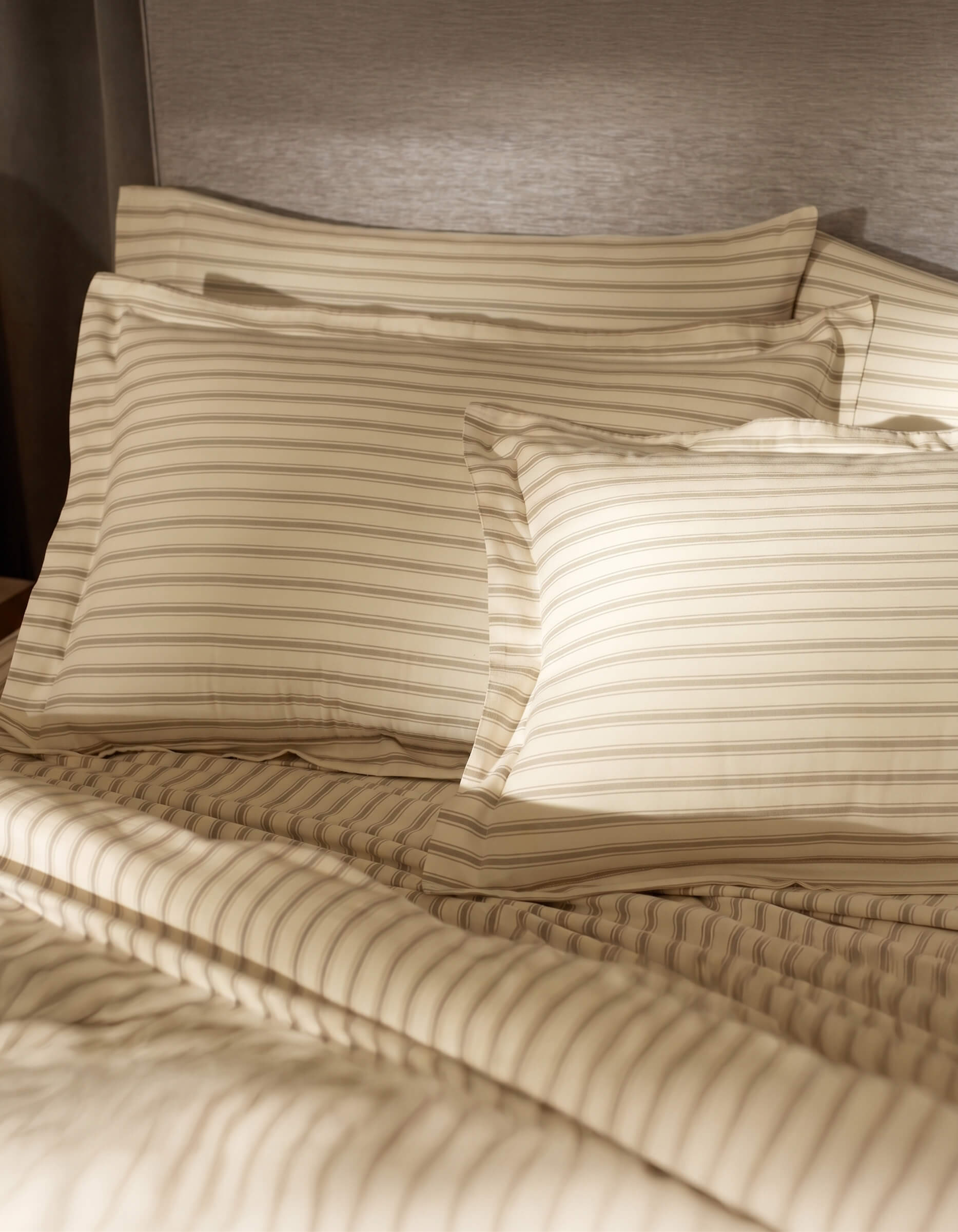 A bed featuring Cozy Earth Soft-Wash Cotton Shams in tan and beige stripes is set against a gray upholstered headboard, softly illuminated by sunlight.
