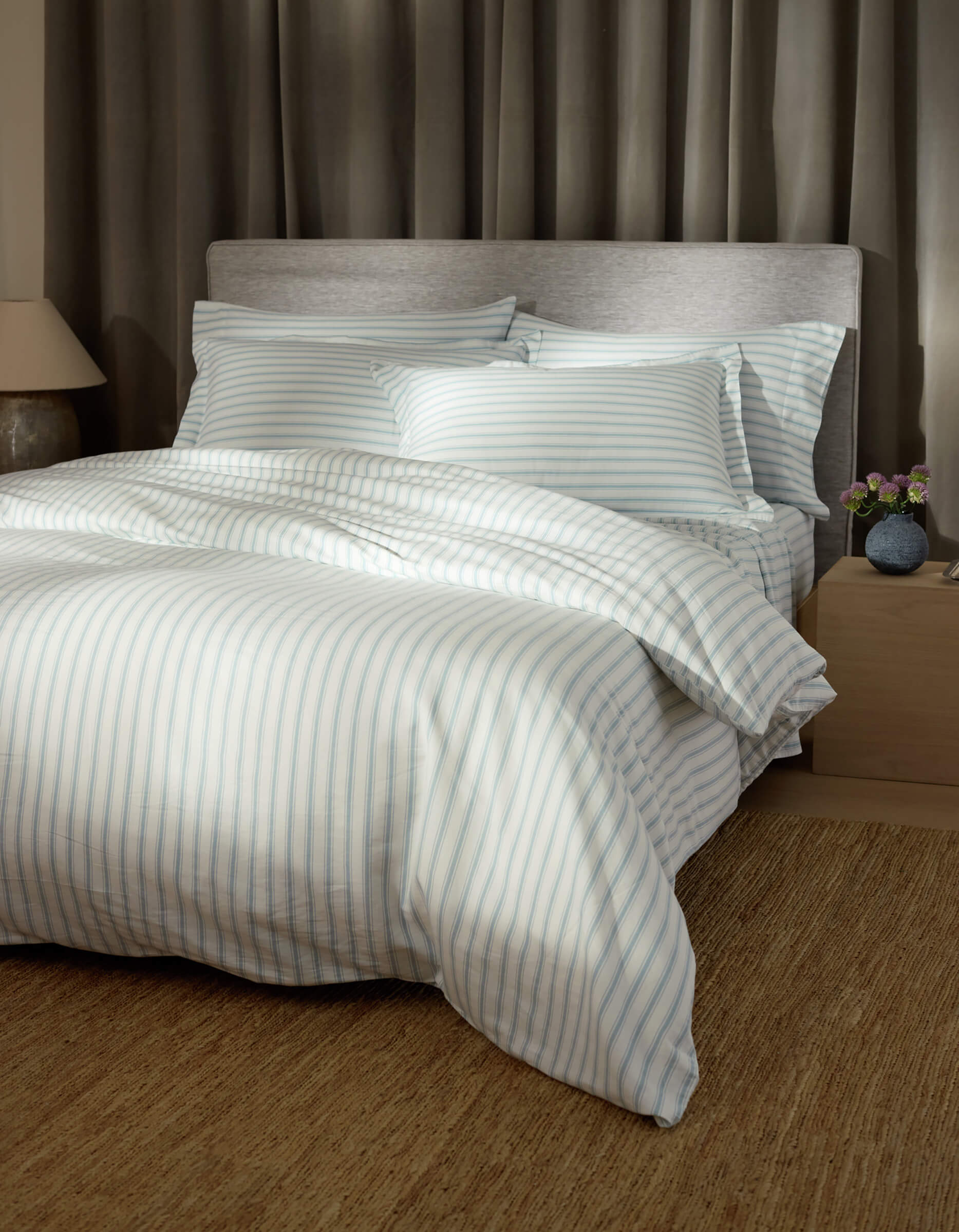 A bed features Cozy Earth Soft-Wash Cotton Shams, light blue and white striped bedding, four pillows, a padded grey headboard, beige walls, a large brown rug, grey curtains, and a nightstand with a lamp and flowers.
