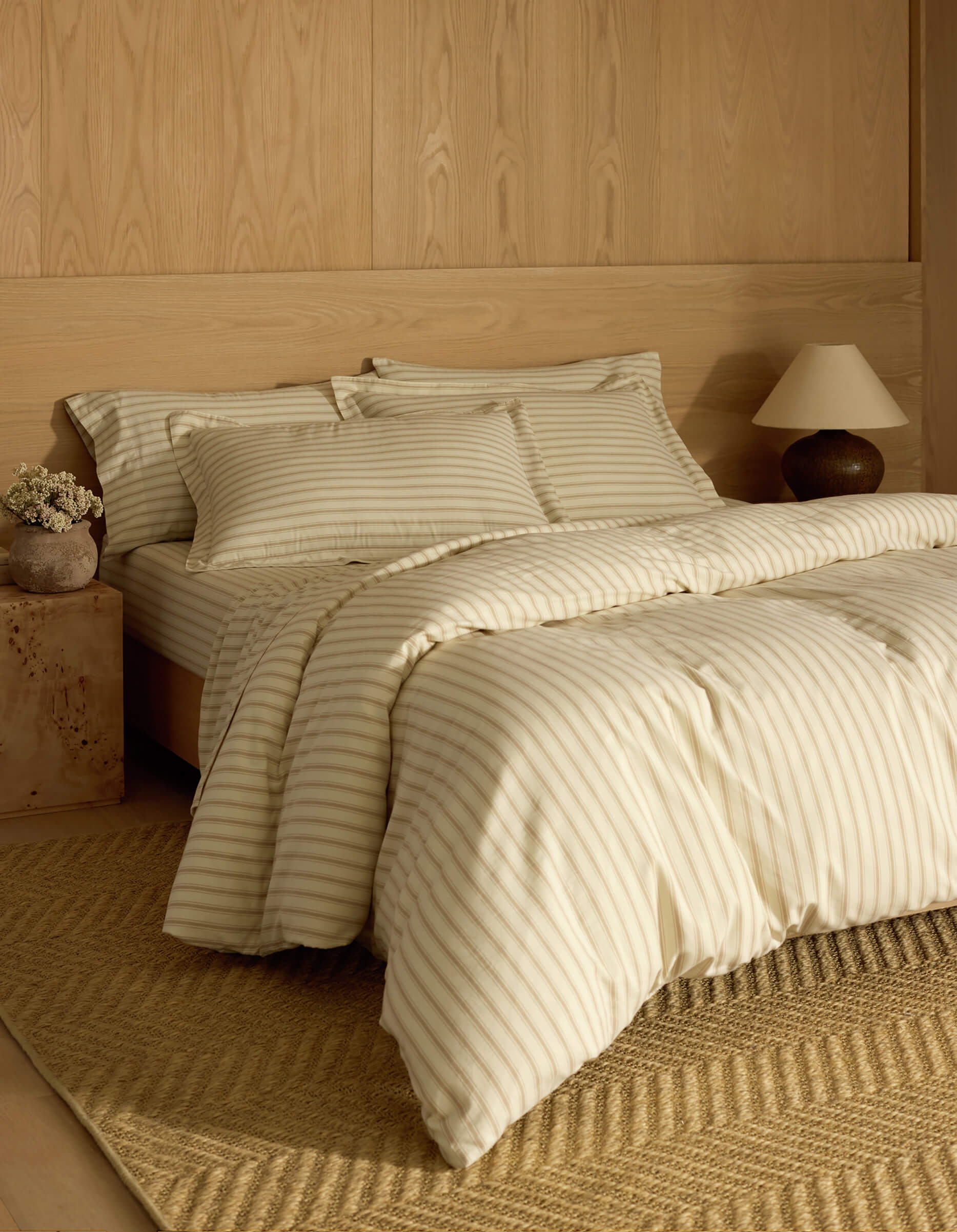 A cozy bedroom features a neatly made bed with Cozy Earth Soft-Wash Cotton Shams in cream and light tan stripes, wood panel walls, a woven rug, small nightstand, ceramic lamp, and a vase of dried flowers.