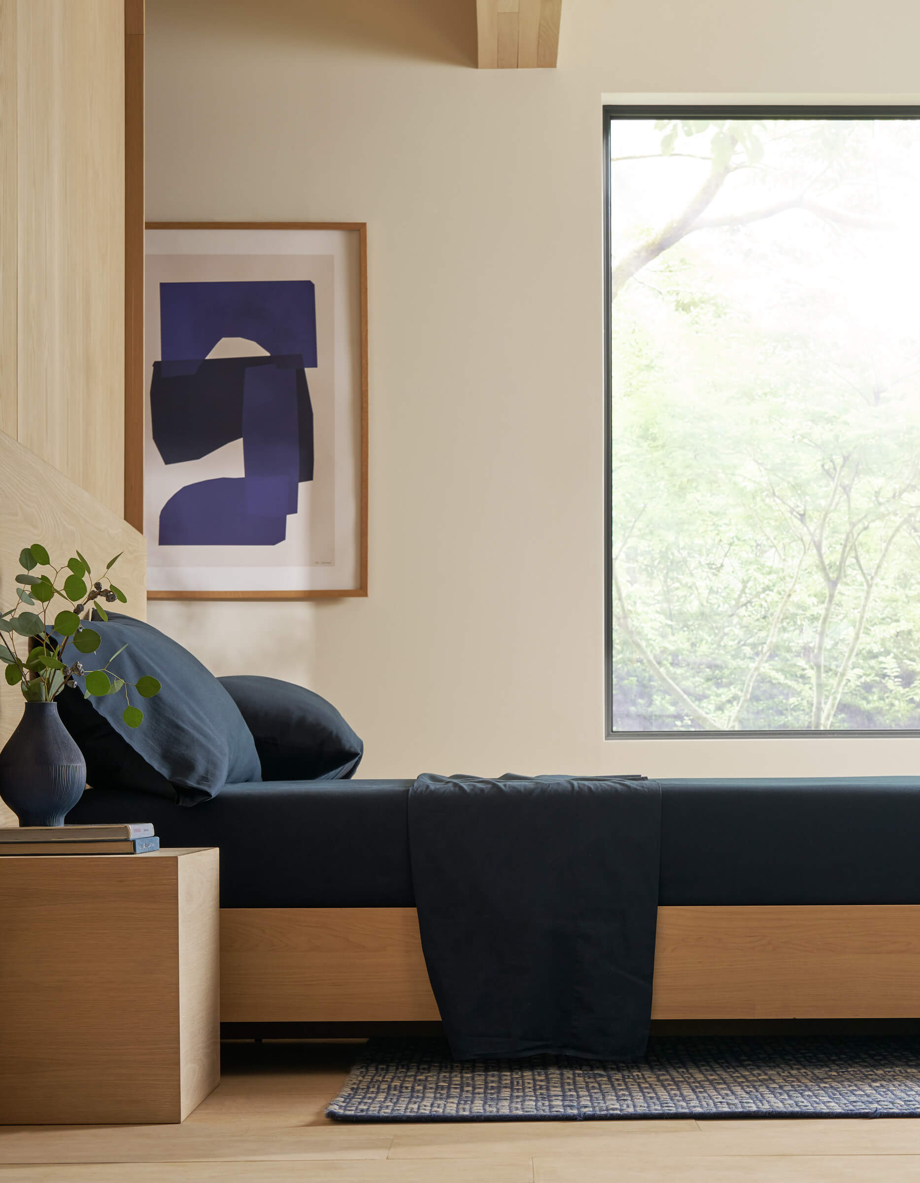 Minimalist bedroom featuring Cozy Earth’s Soft-Wash Cotton Sheet Set in dark blue, a wooden bed frame and nightstand, vase with greenery, abstract art on the wall, and a large window showcasing tree views and natural light.