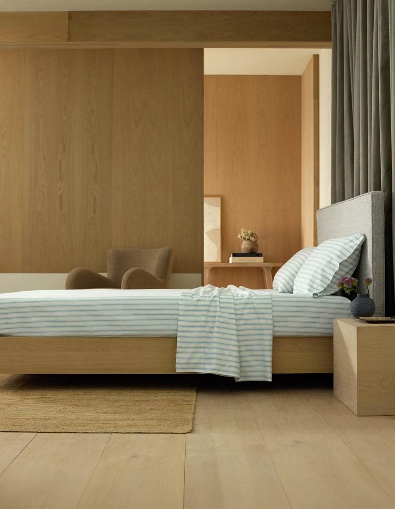 A modern, minimalist bedroom features a low wooden bed dressed in Cozy Earth's Soft-Wash Cotton Sheet Set, striped bedding, a wooden nightstand with a vase, an armchair, and wood-paneled walls and floor bathed in natural light.
