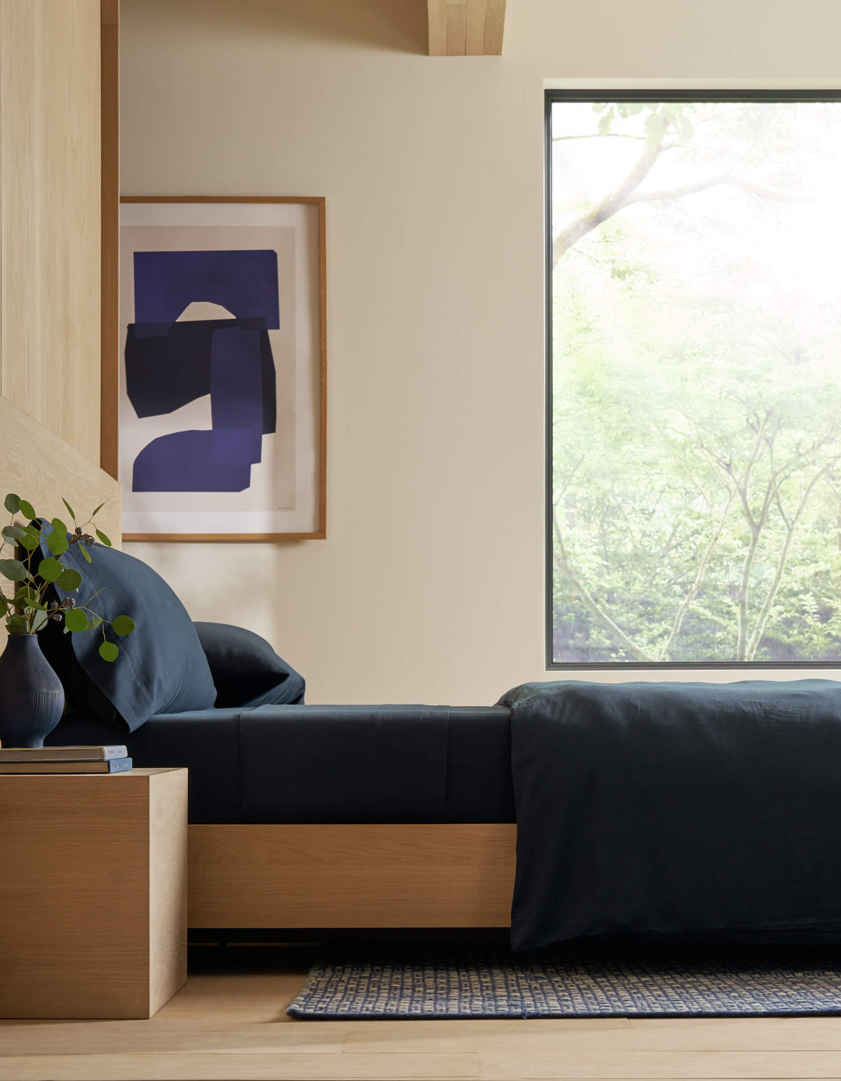 A modern bedroom featuring Cozy Earth's Soft-Wash Cotton Fitted Sheet in dark blue, paired with light wood furniture, a small bedside table adorned with a vase of greenery, abstract blue wall art, and a large window overlooking nature. |Color:Eclipse
