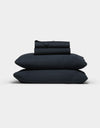 The Cozy Earth Soft-Wash Cotton Sheet Set, in dark navy blue, features two stacked pillows and folded sheets neatly arranged against a plain light gray background.|Color:Eclipse