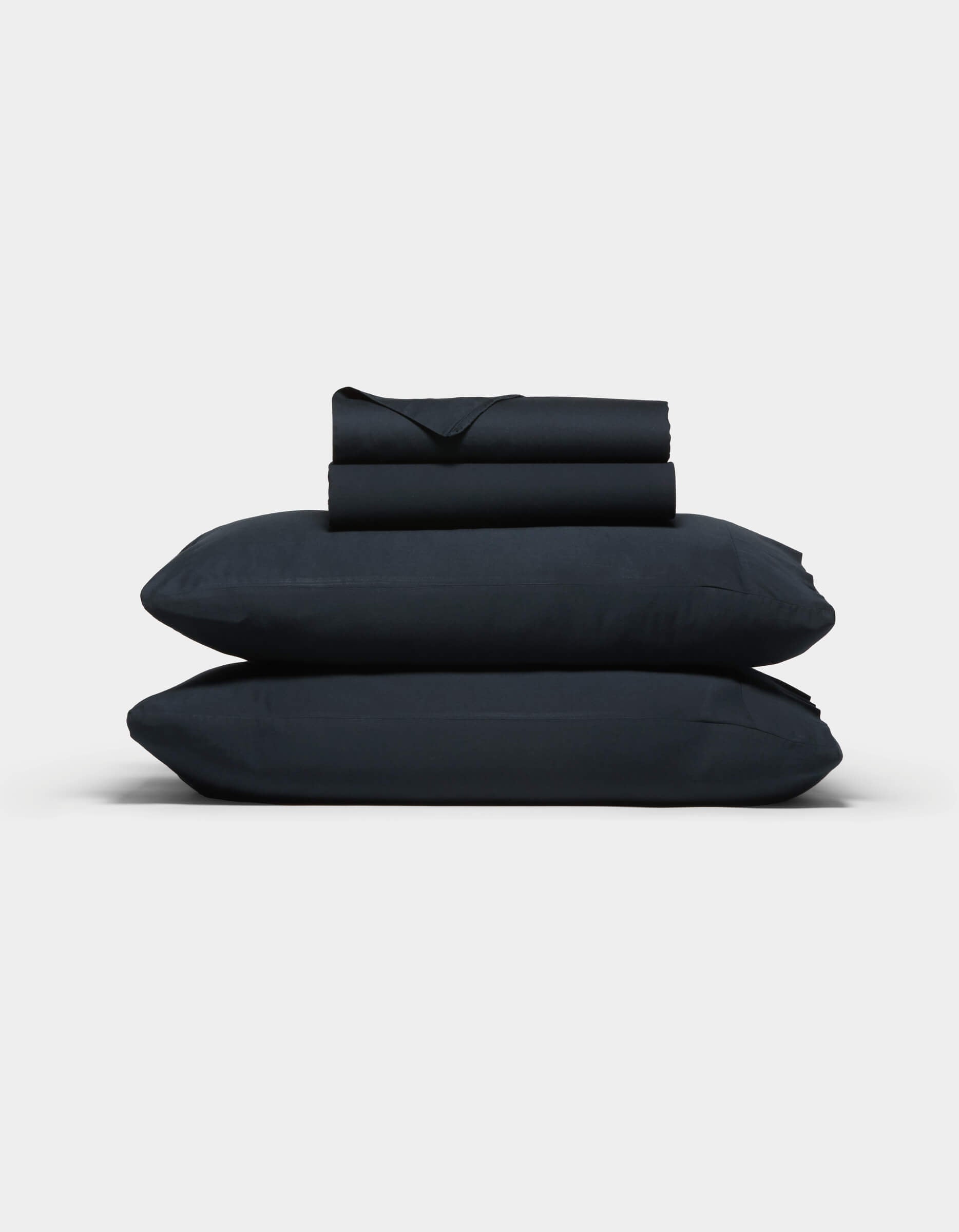 The Cozy Earth Soft-Wash Cotton Sheet Set, in dark navy blue, features two stacked pillows and folded sheets neatly arranged against a plain light gray background.