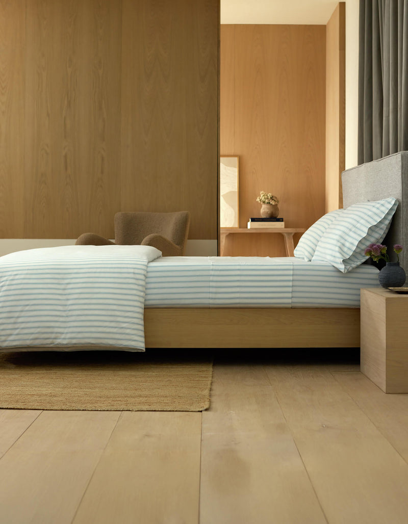 A modern, minimalist bedroom featuring a low wooden bed with Cozy Earth’s Soft-Wash Cotton Fitted Sheet, striped bedding, a beige rug, gray headboard, wooden nightstand with small vases, armchair, and light wood paneling.