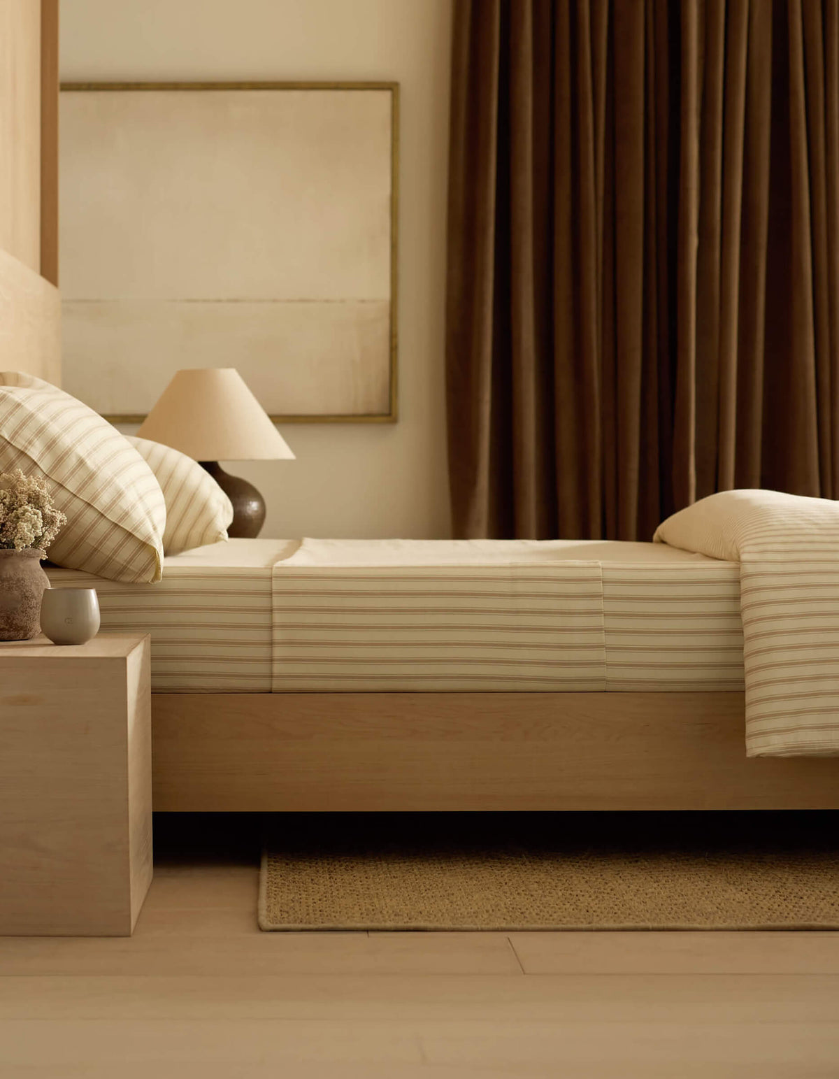 A neatly made bed with Cozy Earth’s Soft-Wash Cotton Sheet Set in beige and white stripes sits beside a wooden nightstand holding a lamp, vase of flowers, and cup in a softly lit, neutral-toned bedroom.|Color:Coconut and Sandalwood Stripe