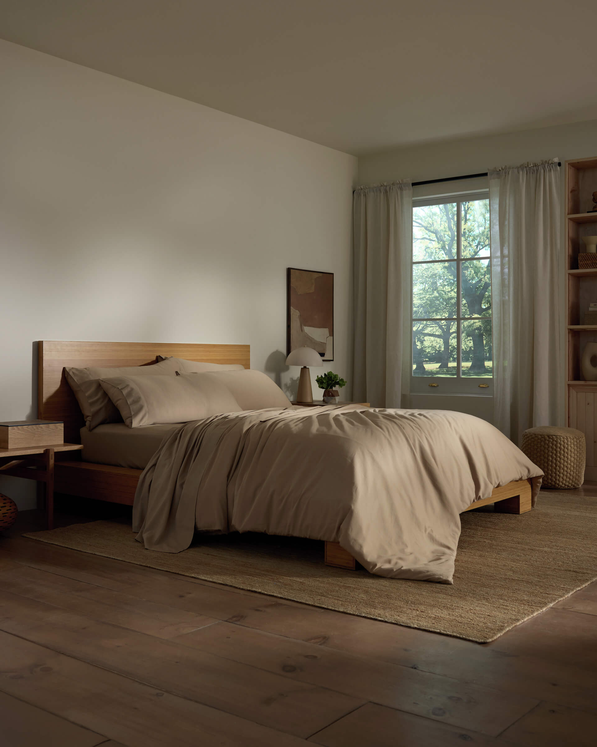 A cozy bedroom features a wooden bed frame with the Soft-Wash Cotton Bedding Core Bundle by Cozy Earth, beige bedding, a nightstand and lamp, neutral décor, large window with white curtains, wooden floors, and abundant natural light.