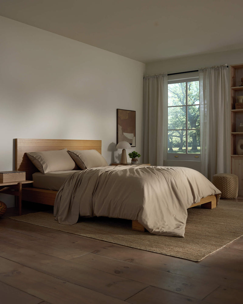 A cozy bedroom featuring a wooden bed frame with Cozy Earth's Soft-Wash Cotton Bedding Deluxe Bundle in beige, a nightstand with a lamp and plant, sheer curtains letting in natural light, and wooden floors topped by a woven rug.